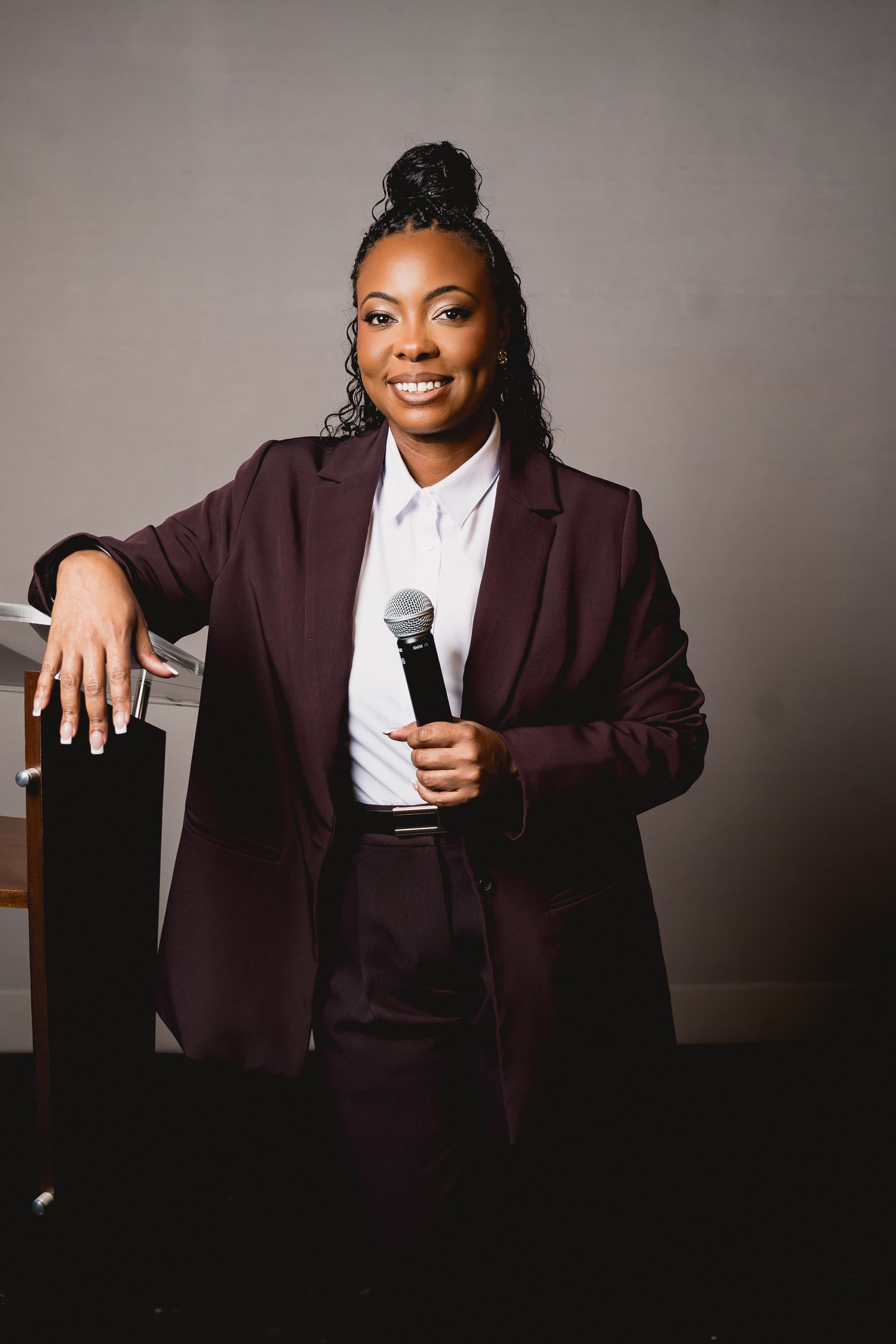 Professional speaker in burgundy suit holding microphone against gray background.