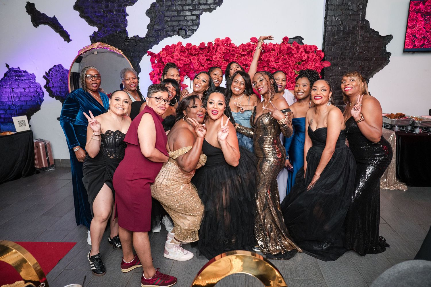A joyful group of women in elegant black, gold, and burgundy dresses pose together at an upscale event with a red floral wall behind them.