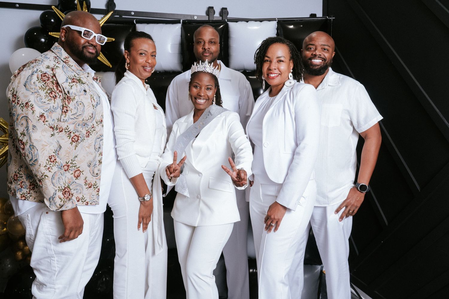 Six smiling people in coordinated white outfits pose together against a dark backdrop, one wearing a floral jacket and sunglasses.