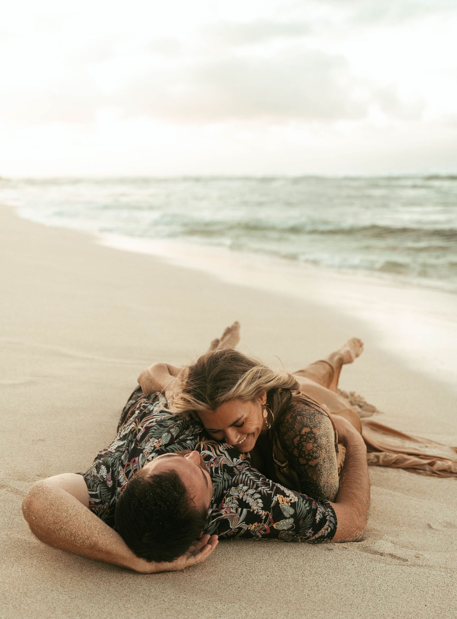 A person in a floral dress lies serenely on a sandy beach as waves roll in during sunset.