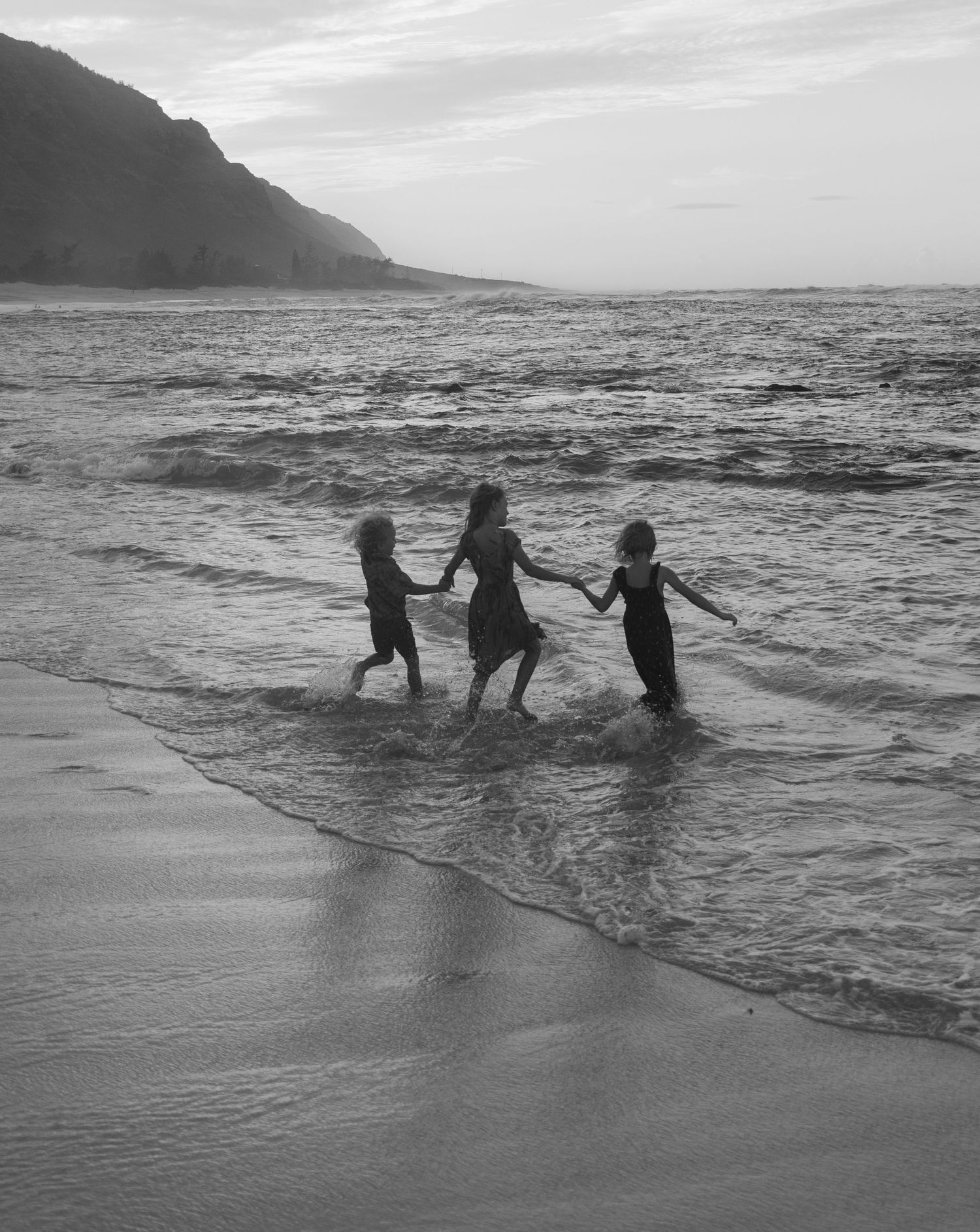 Three silhouettes dance joyfully in the ocean waves at sunset with mountains in the background in black and white.