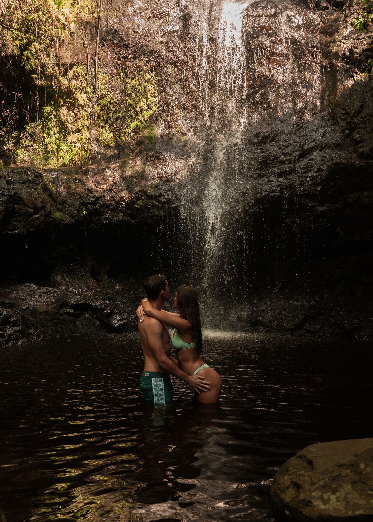 A swimmer dives into a dark cenote pool beneath a natural rock formation with sunlight streaming through the cave opening.