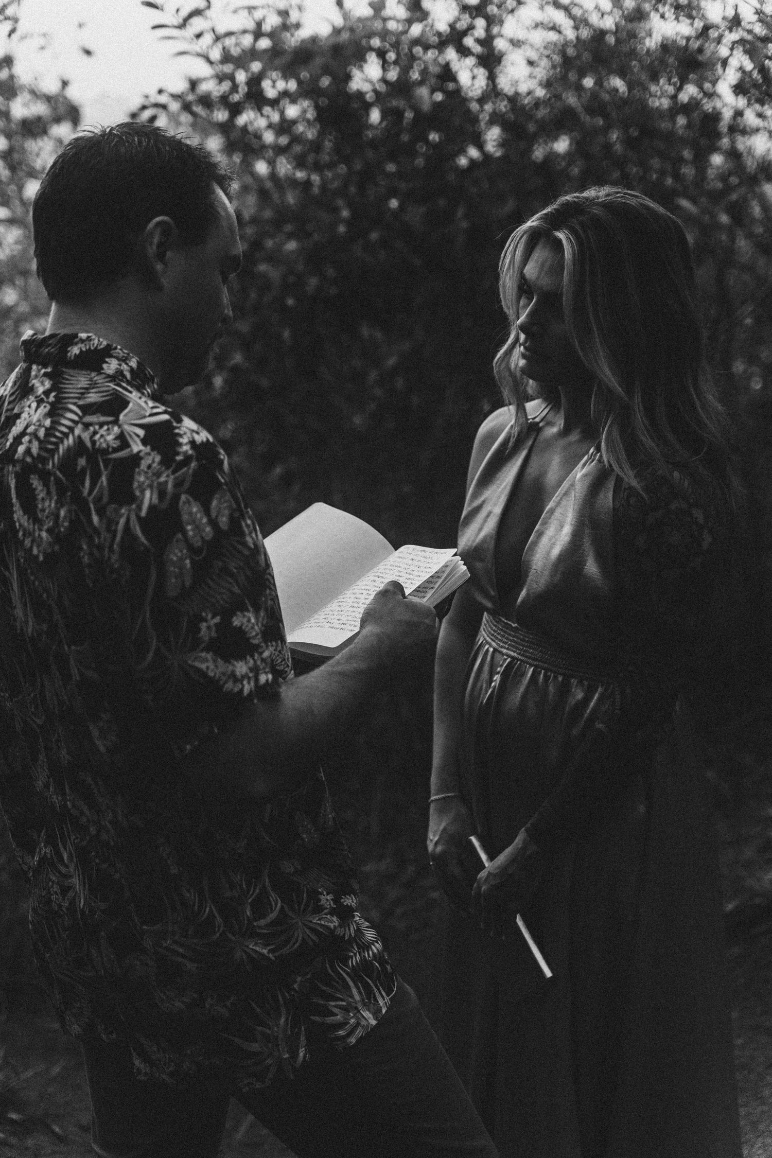 A black and white photo of a couple sharing a tender moment while reading together outdoors in nature.