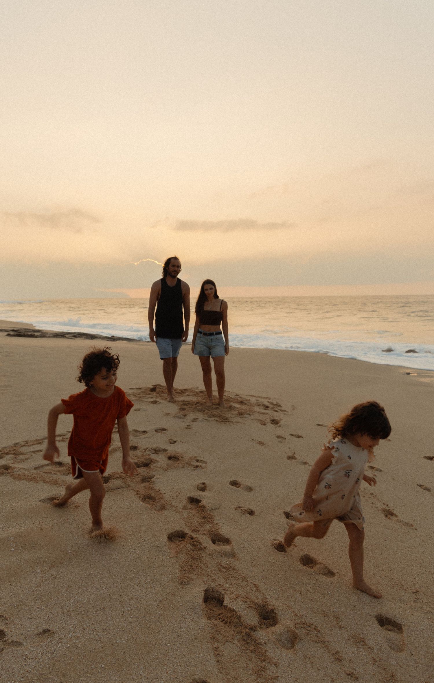Two children run playfully on a beach at sunset while two figures watch from behind on the golden sand.