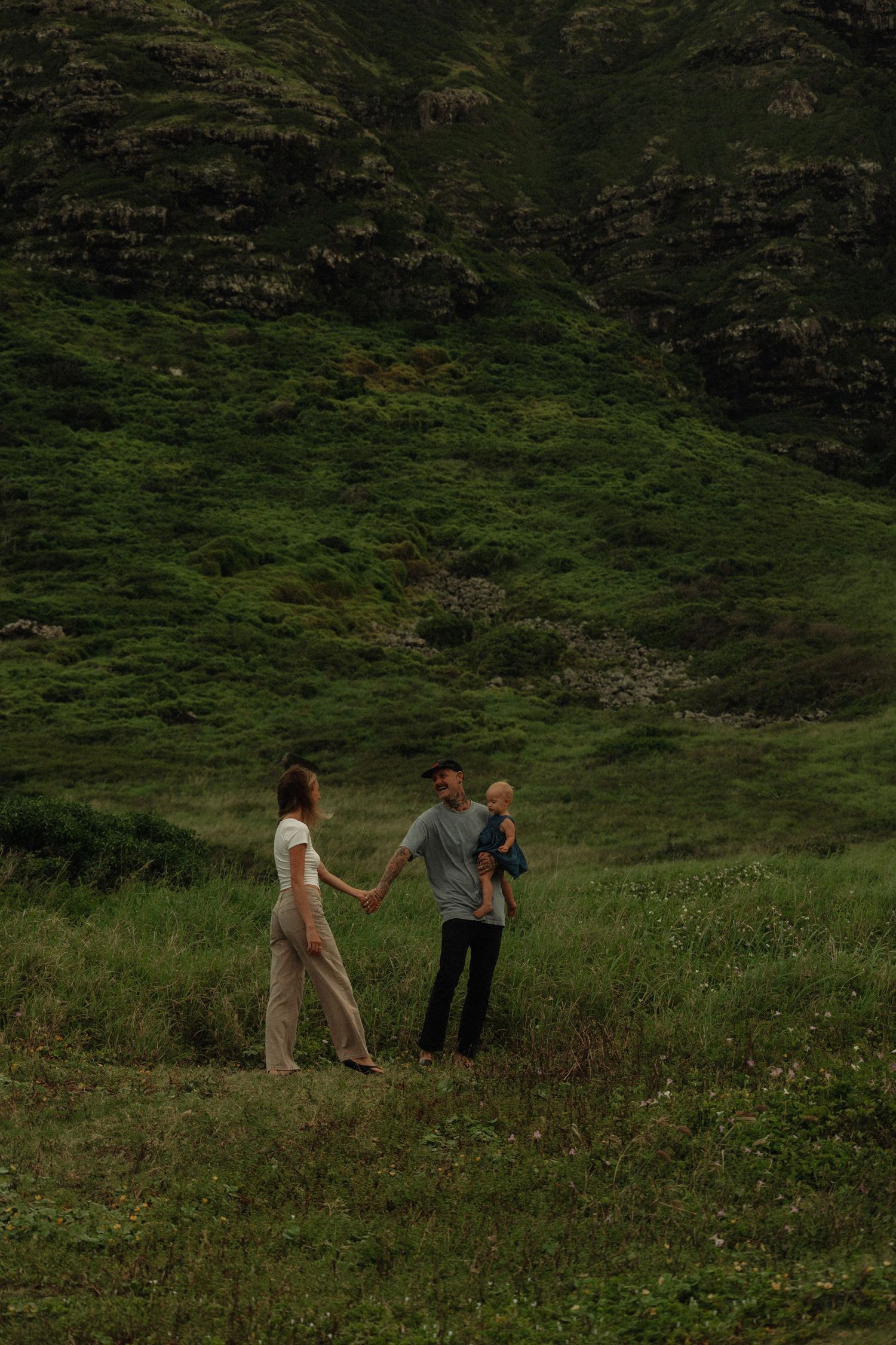 A couple holds hands in a lush green meadow, man carrying a baby, with rolling hills in the background.