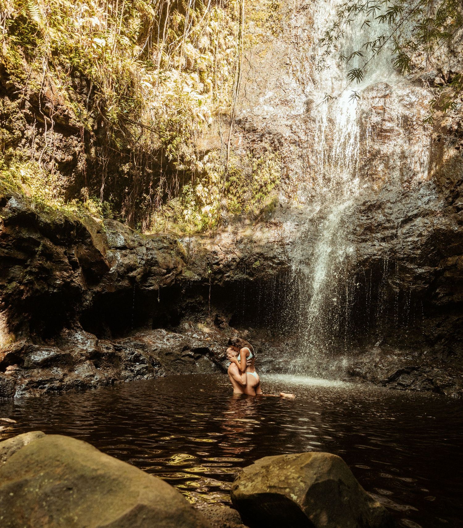 A scenic waterfall cascades into a tranquil natural pool surrounded by lush tropical vegetation and rock formations.