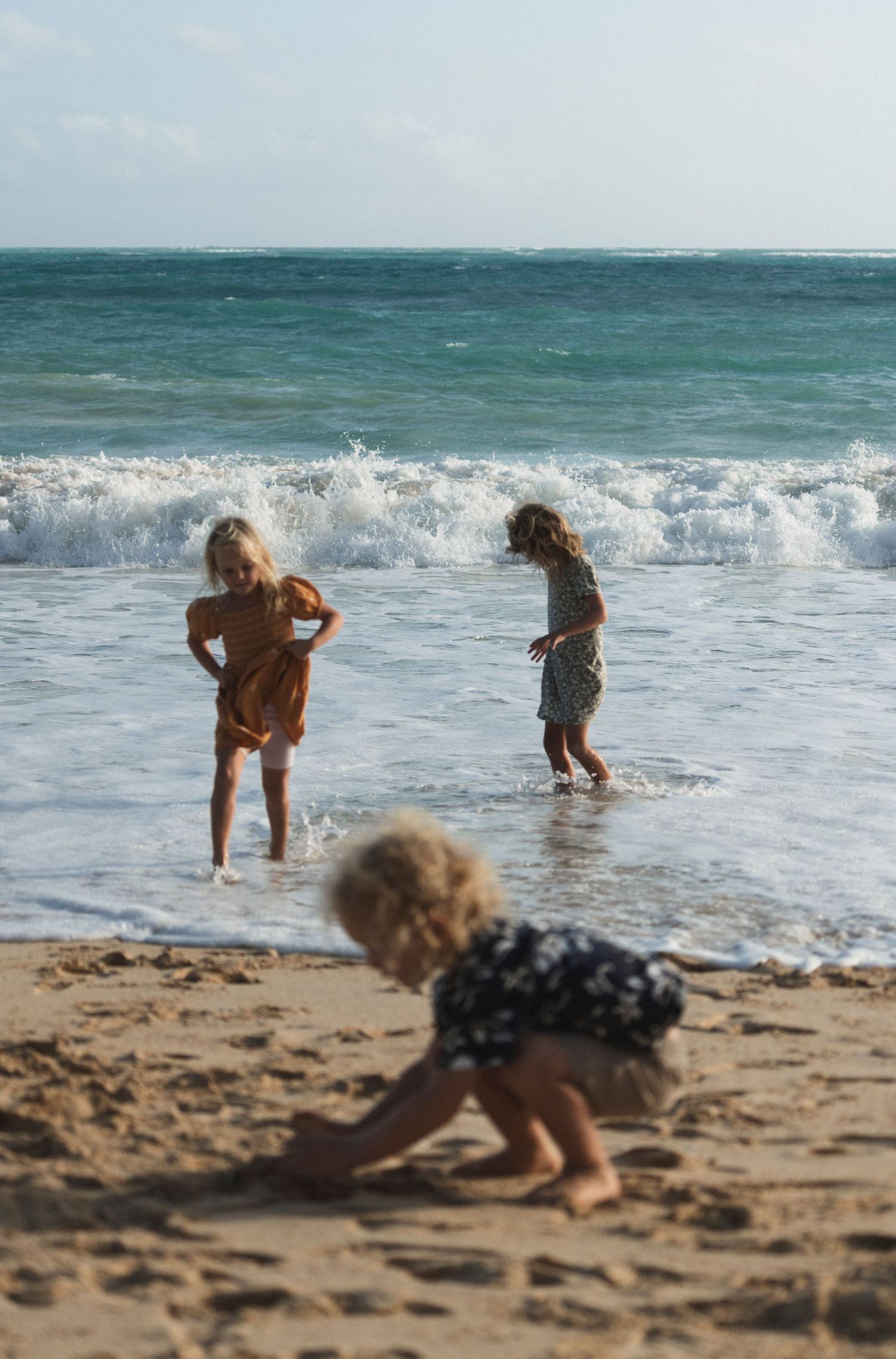 Three children play on a sandy beach as turquoise waves crash on the shore during a sunny day.