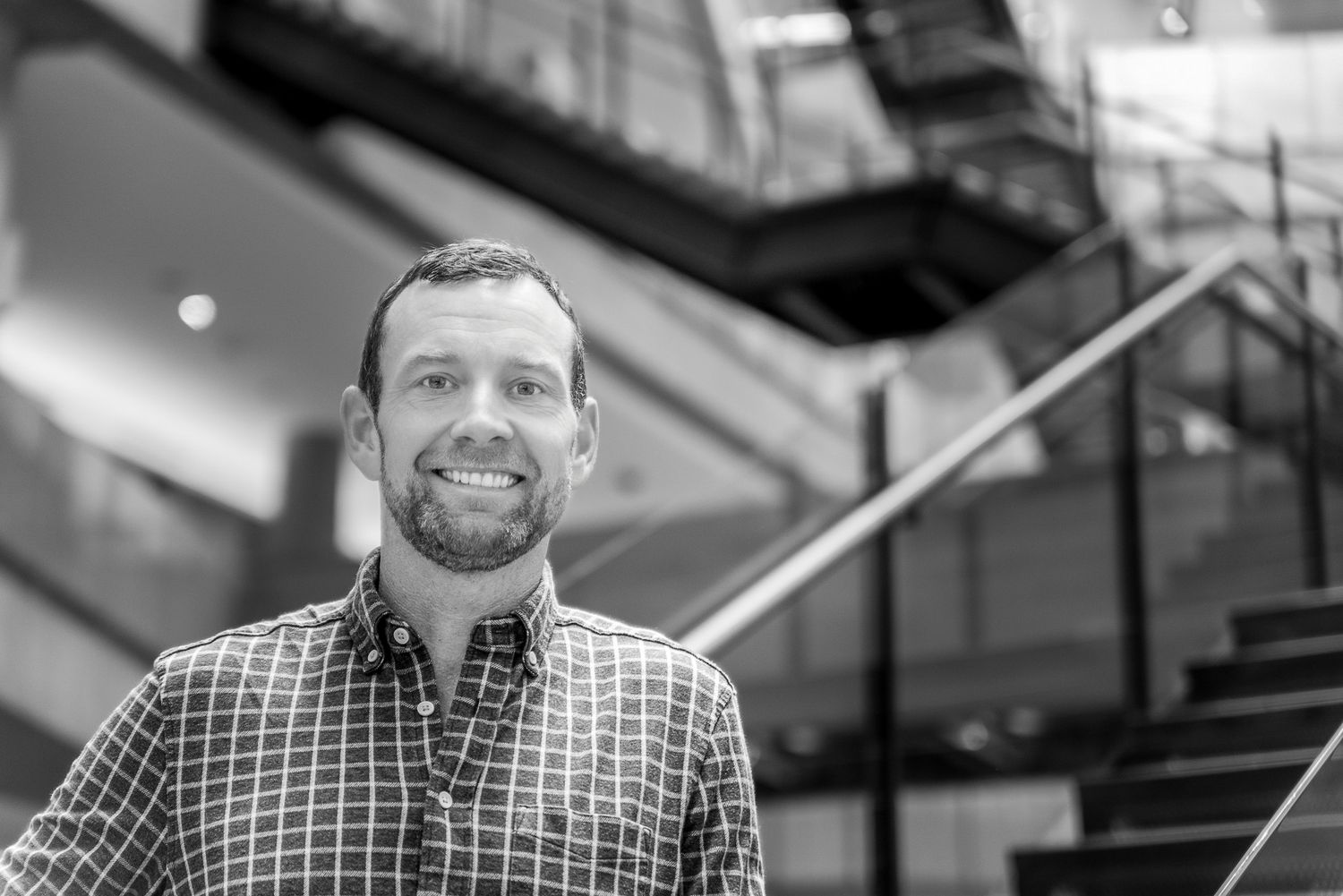 Black and white portrait of someone smiling in a plaid button-down shirt with stairs and railing in the background.
