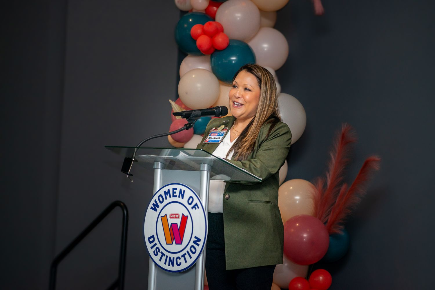 Presenter speaking at podium decorated with balloons and company logo during event.