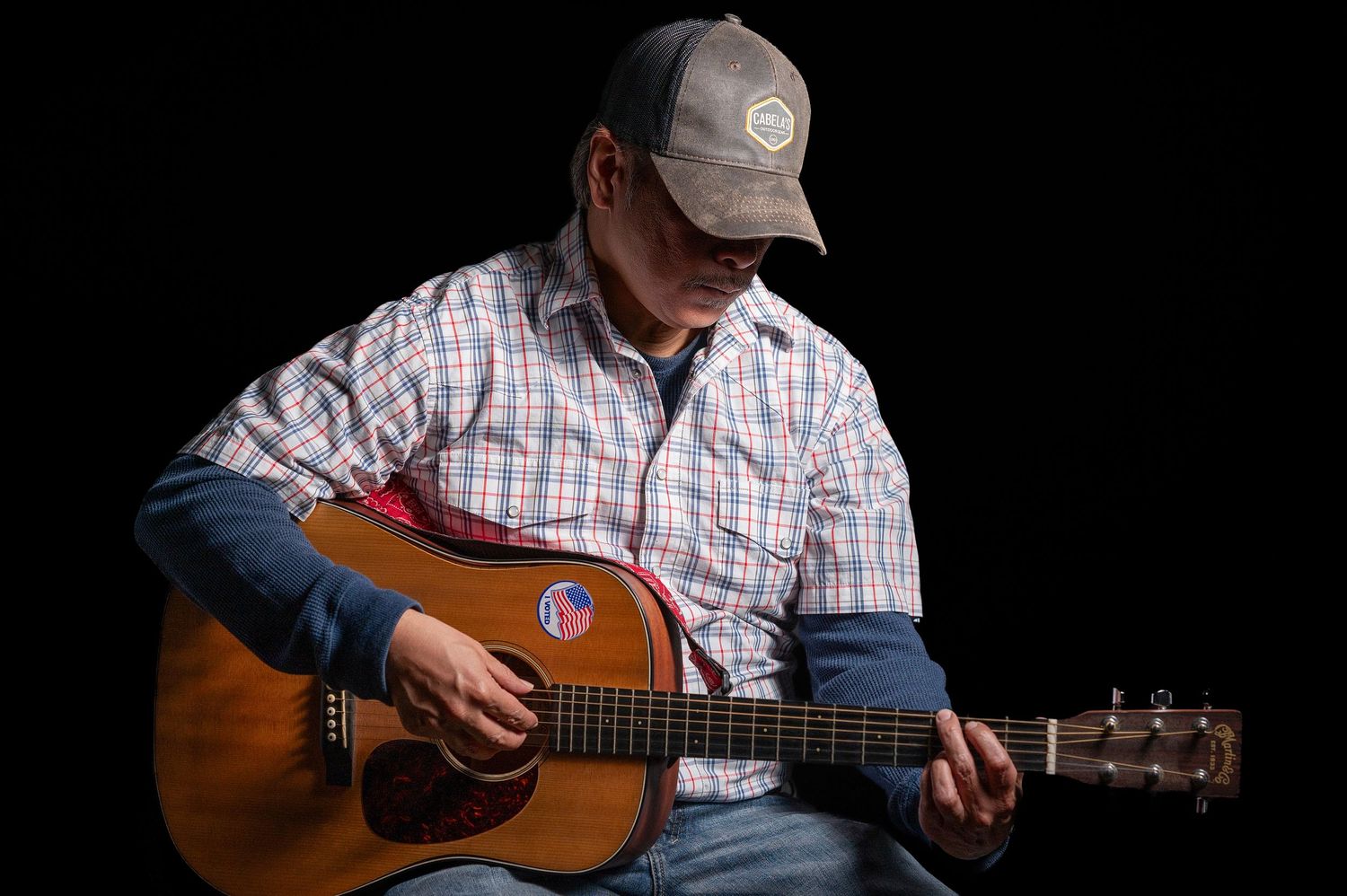 Musician playing acoustic guitar against black background in casual attire.
