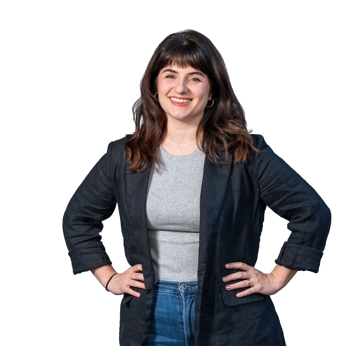 Professional portrait against white background showing someone in a black blazer and gray shirt with hands on hips.