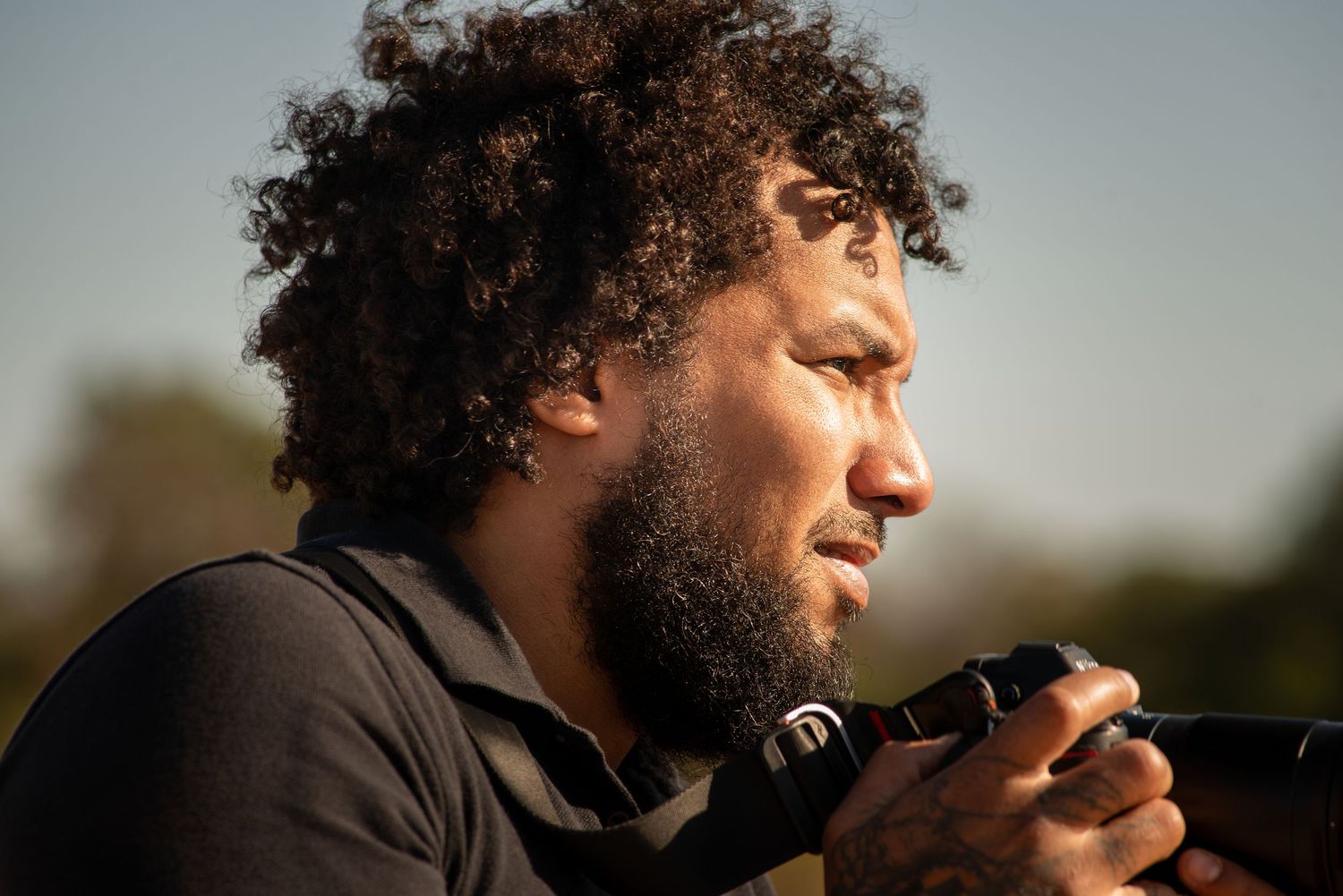 A photographer with curly hair and a beard looks through a camera lens in warm outdoor lighting.