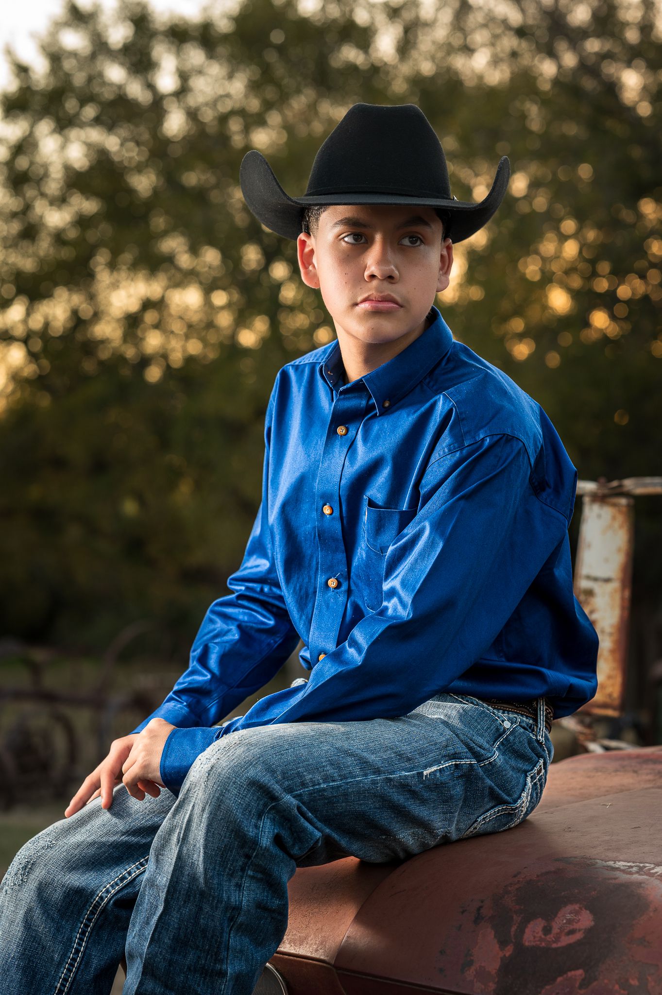 Young child in blue western shirt and cowboy hat sits on rustic wooden fence at sunset.