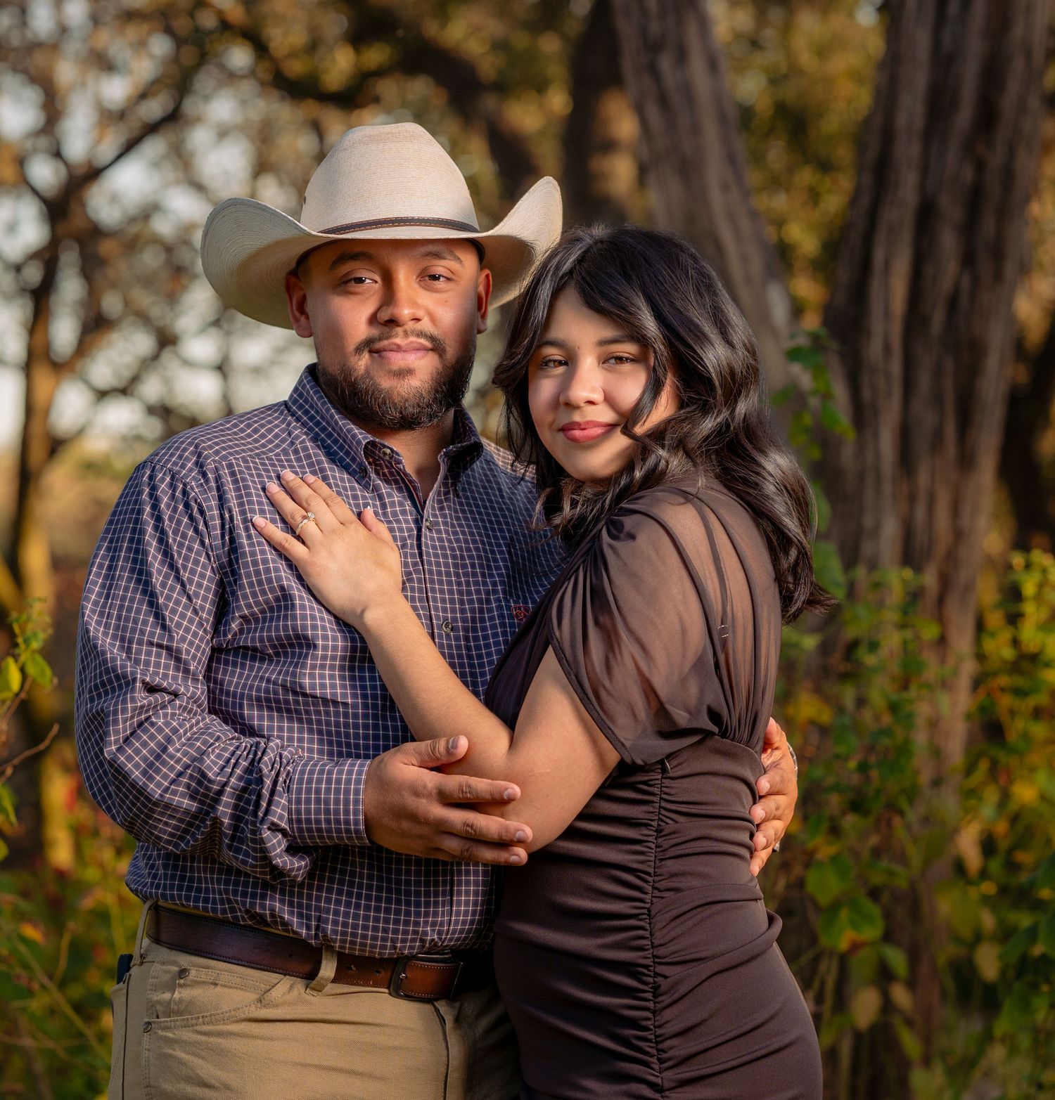 A couple embraces outdoors during golden hour with one wearing a cowboy hat and plaid shirt.