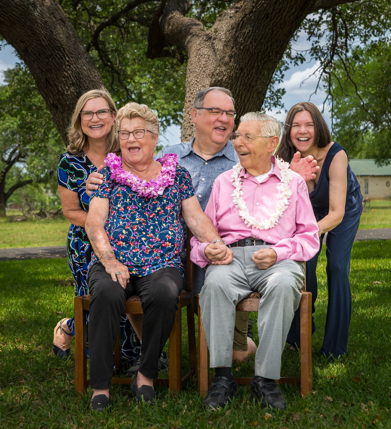 Multi-generational family group seated on chairs under tree for outdoor portrait.