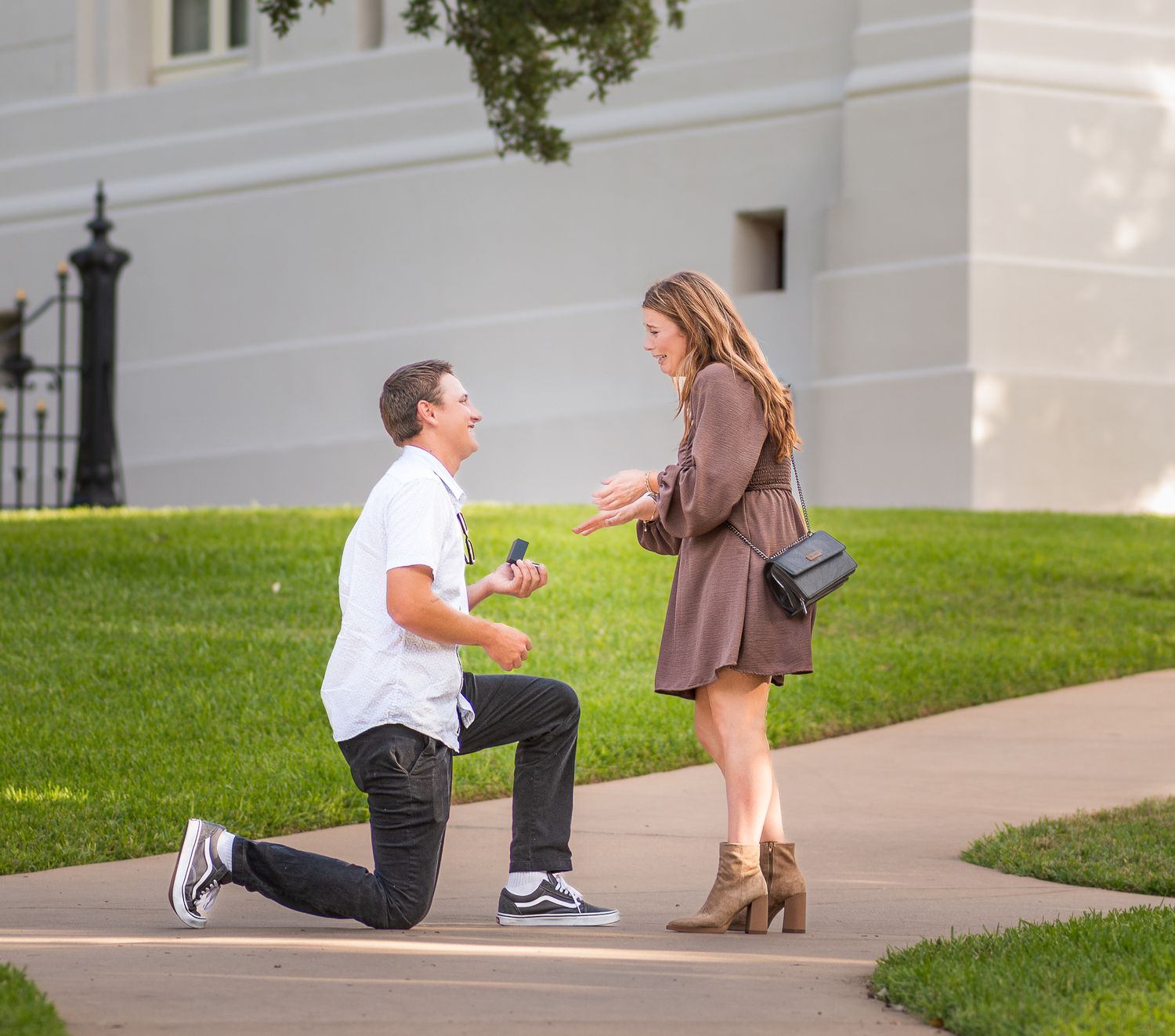 A romantic proposal sequence showing a couple on a grassy slope outside a white building as someone kneels down.