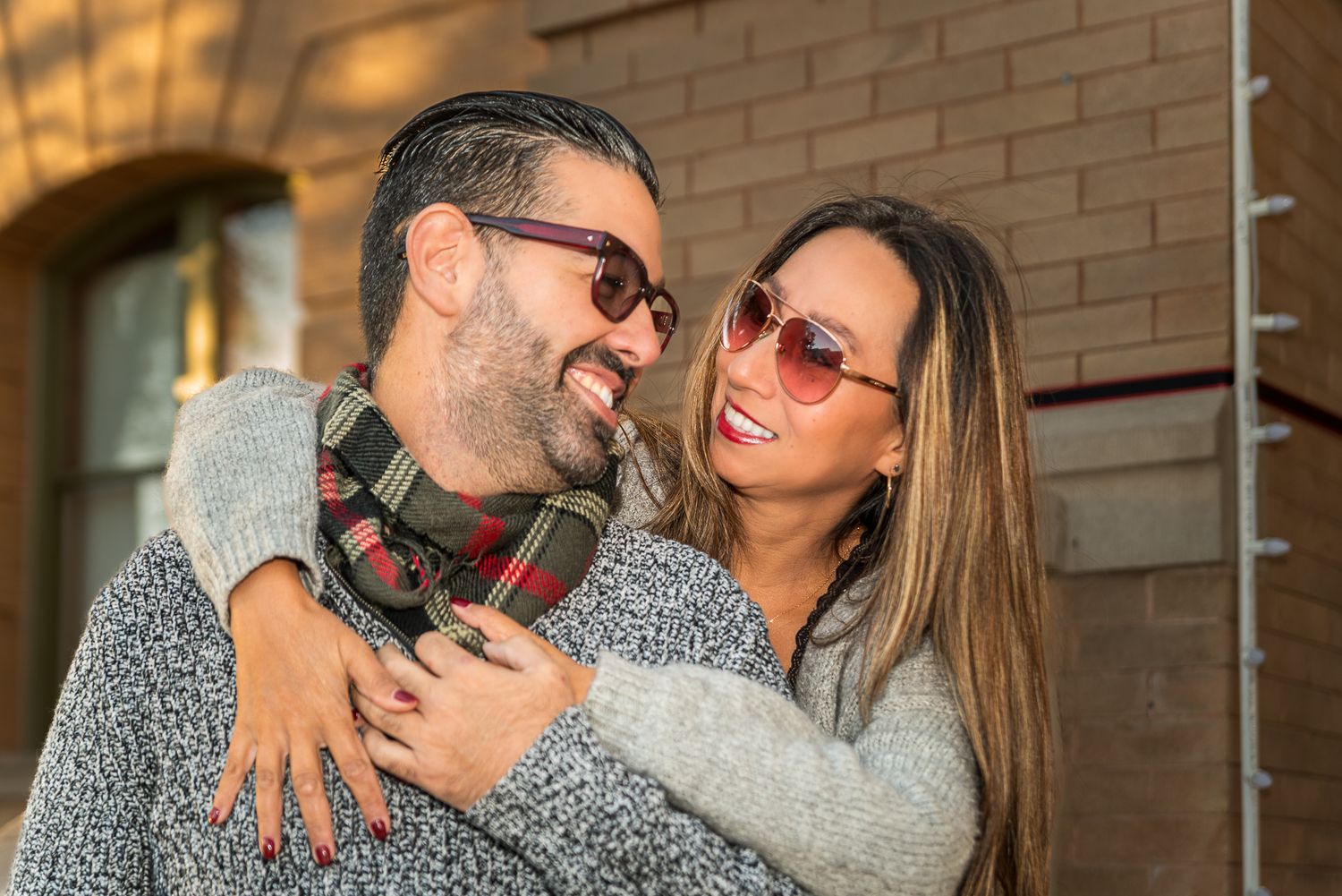Couple embracing and laughing together against brick wall during casual photoshoot.