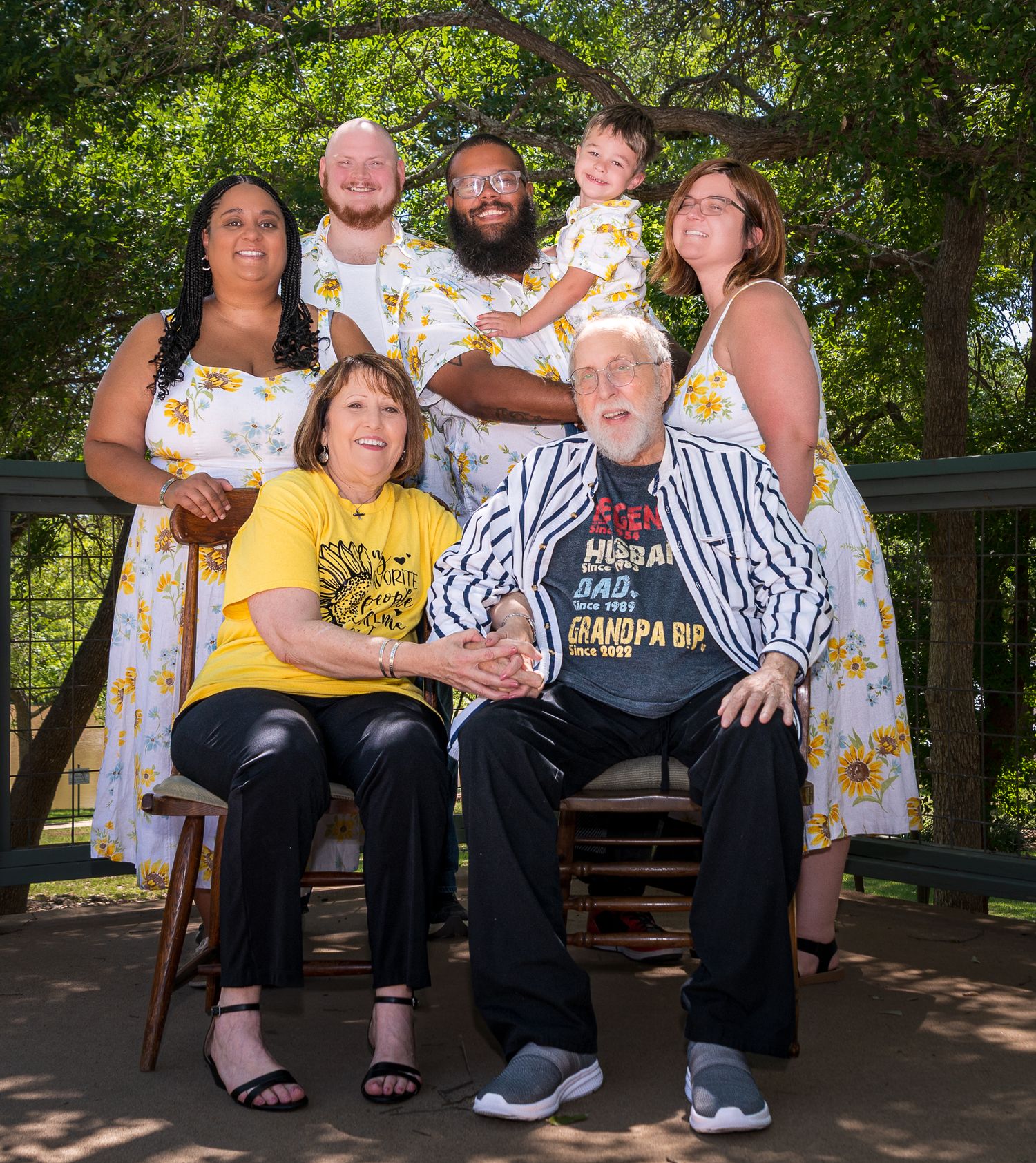 Casual family portrait session on wooden bench in shaded outdoor setting.