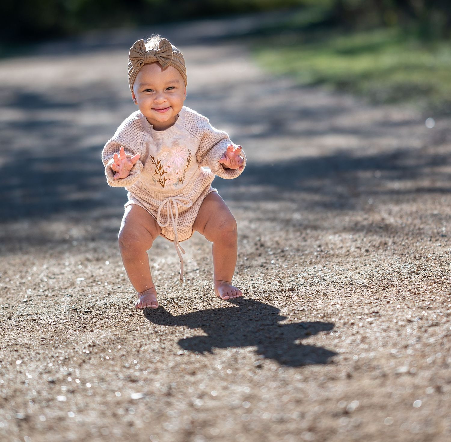 A toddler in a white dress squats down to play on a gravel path while casting a shadow in bright sunlight.