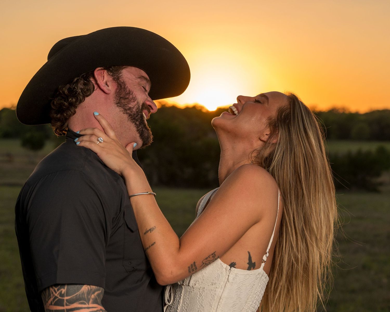 Couple sharing a romantic moment at golden sunset, man in cowboy hat with tattoos, woman in white dress laughing joyfully.