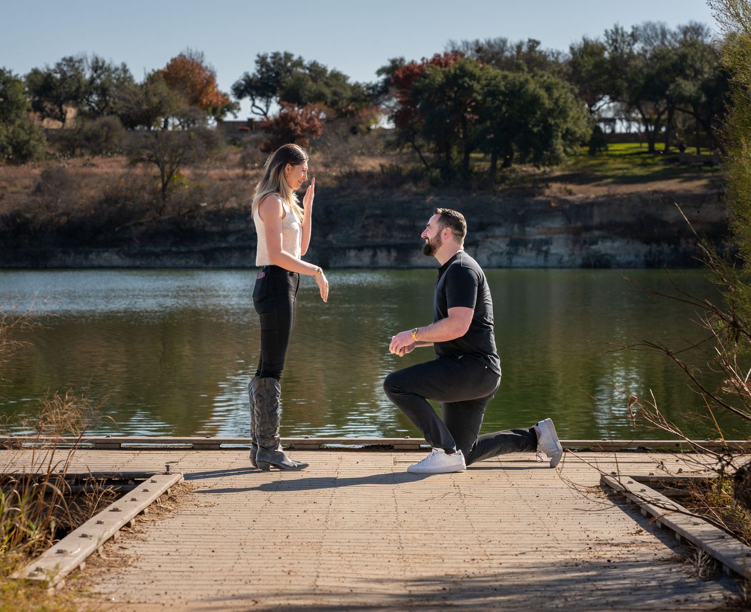 A romantic proposal scene unfolds on a wooden dock overlooking a serene lake surrounded by autumn trees.