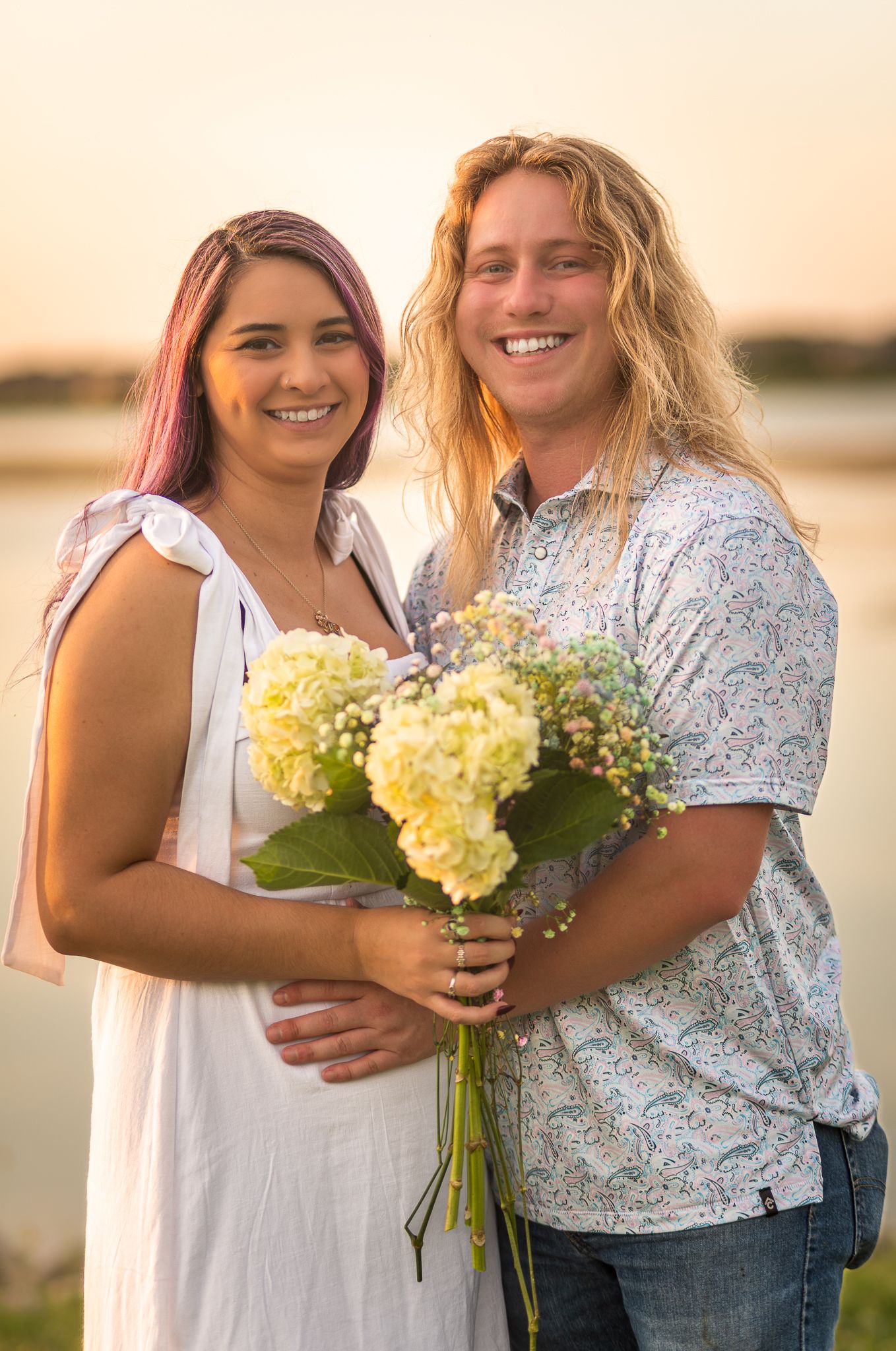 Two friends hold yellow hydrangea flowers while posing together at sunset by the water.