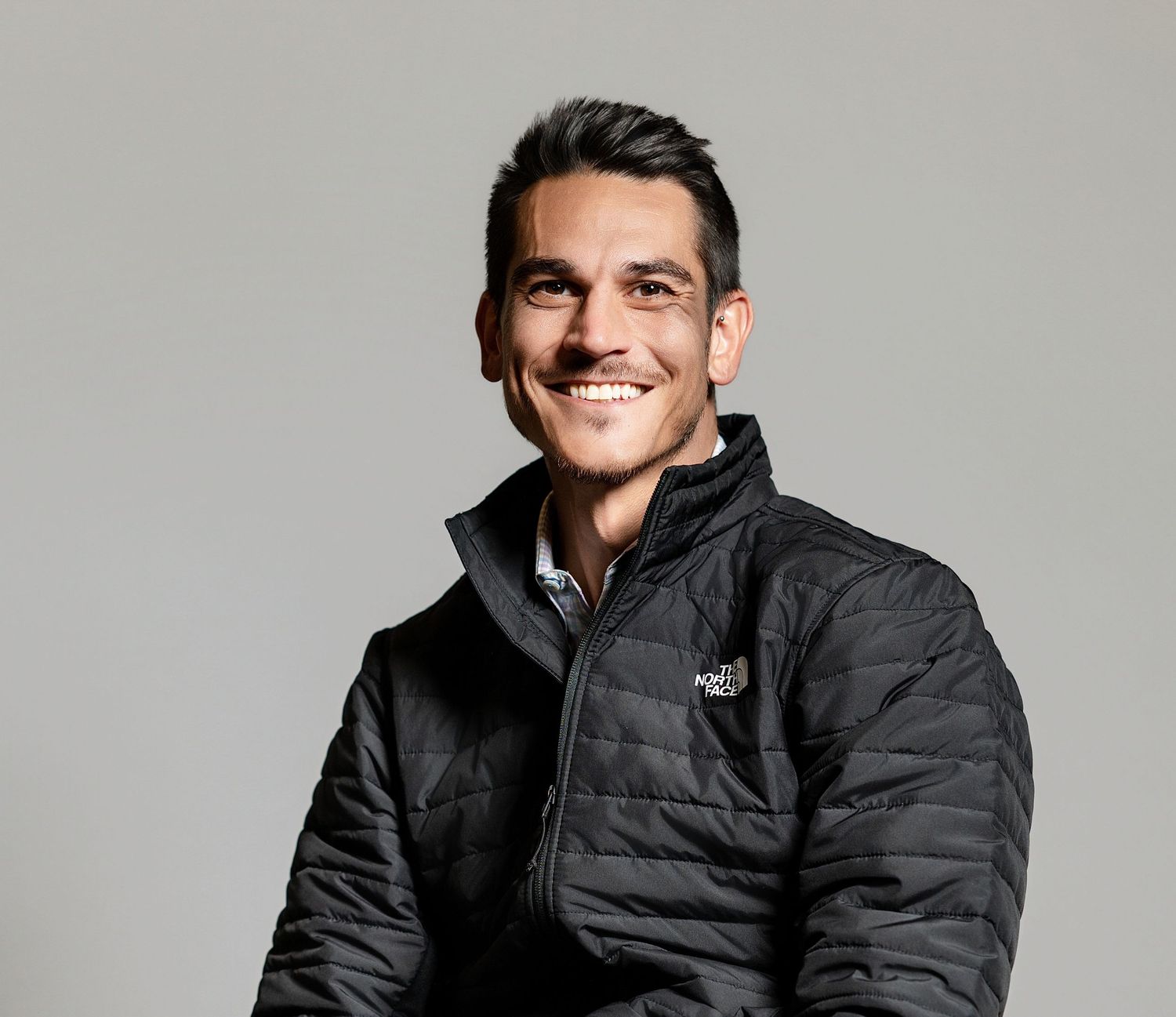 A person wearing a black puffer jacket smiles warmly while posing against a gray background.