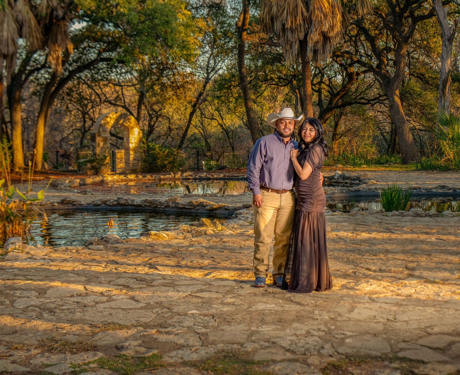 A couple stands together in a golden sunset at a scenic outdoor location, surrounded by trees and natural light.