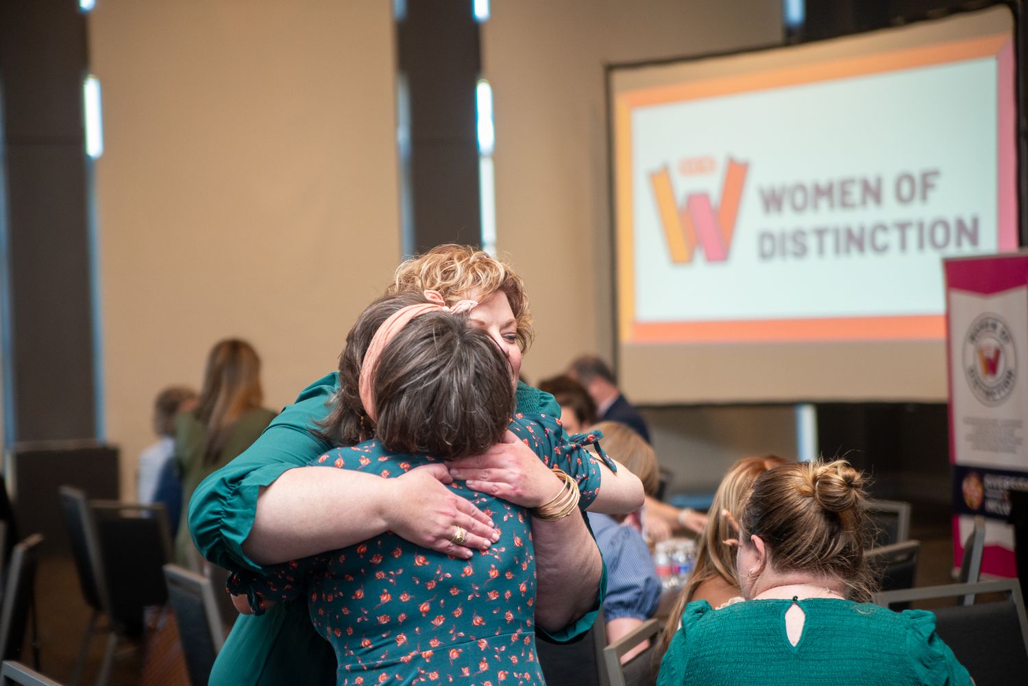 Emotional group hug at Women of Distinction event with projection screen visible.