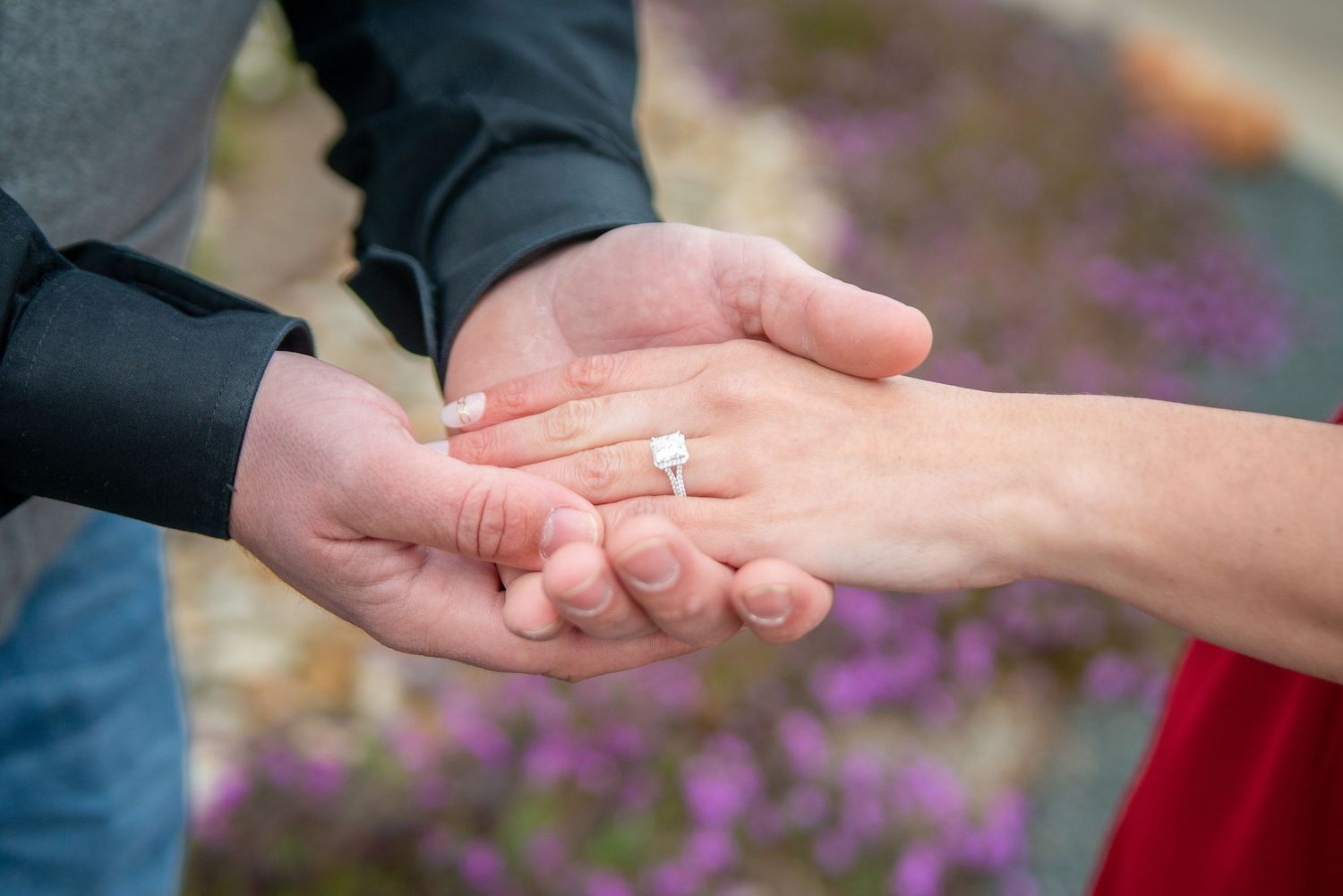 Close up of hands showing engagement ring with purple flowers in background.