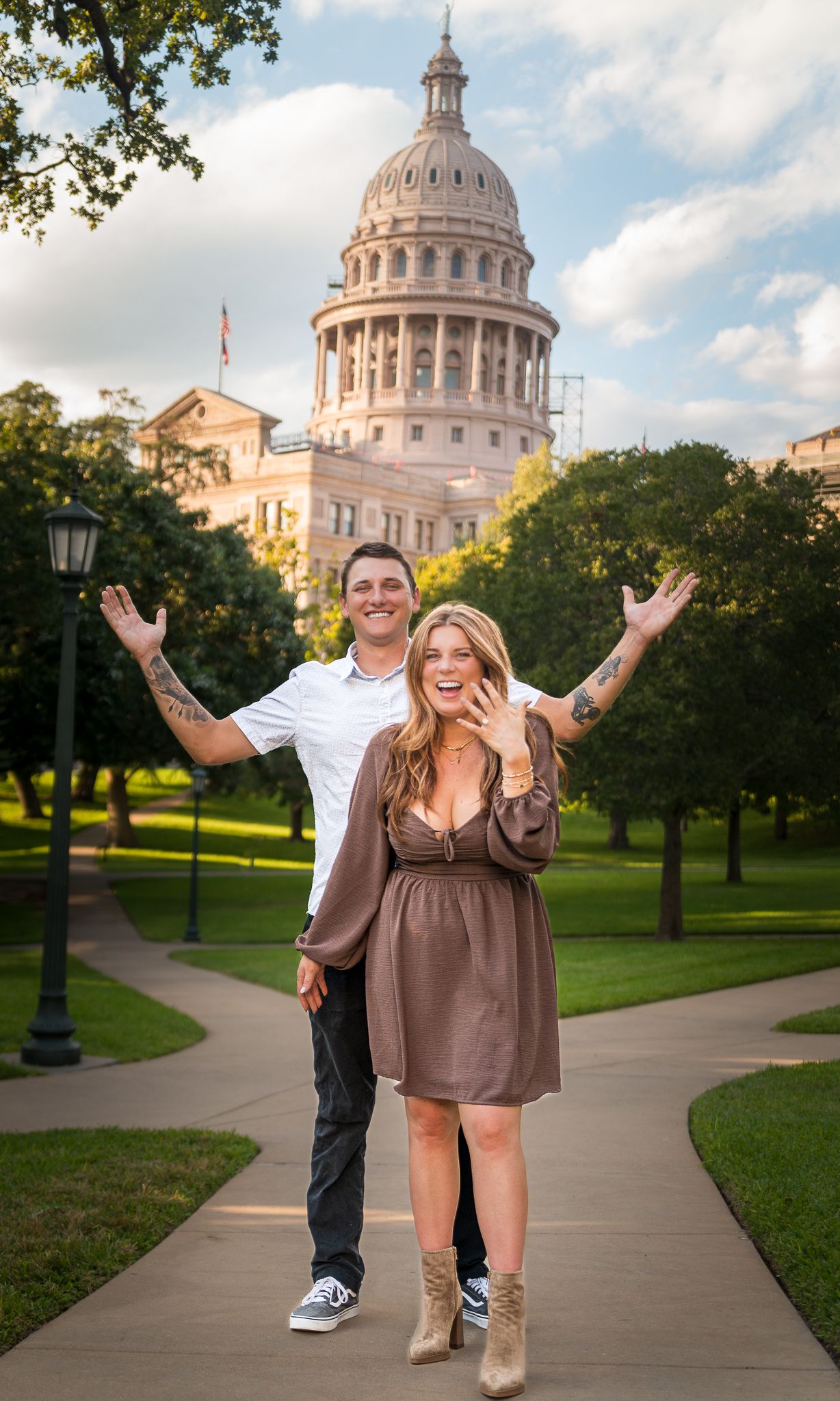 A couple poses cheerfully on a path in front of the Texas State Capitol building in Austin on a sunny day.