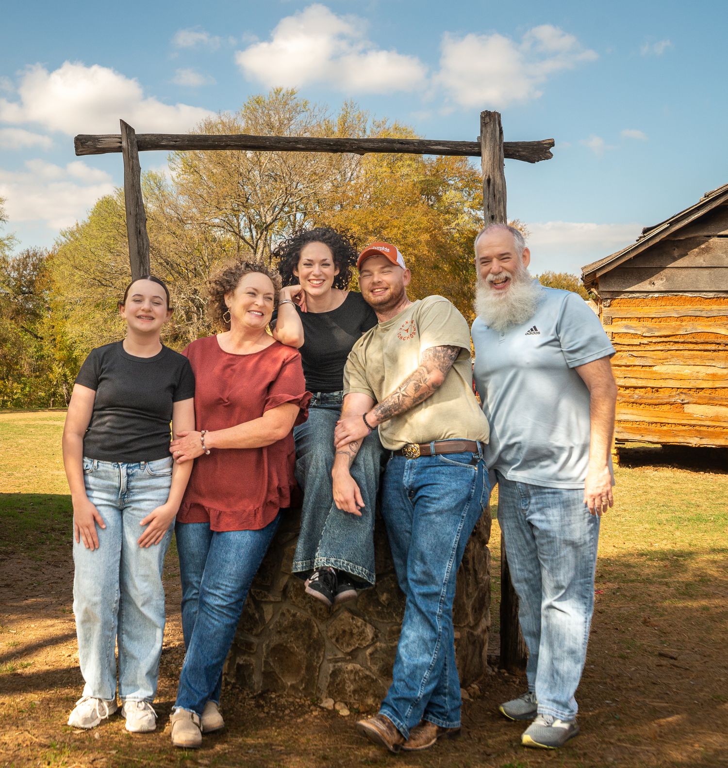 Group of people sitting and standing together on a wooden bench against rustic log cabin walls in autumn scenery.