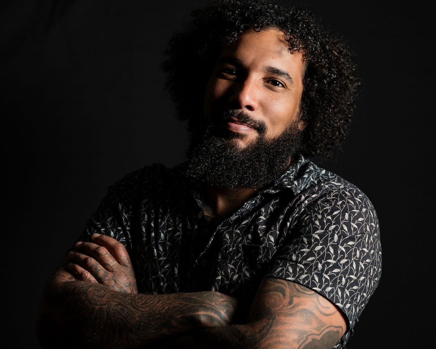 Portrait of person with curly hair and beard wearing patterned shirt against dark background with arms crossed.