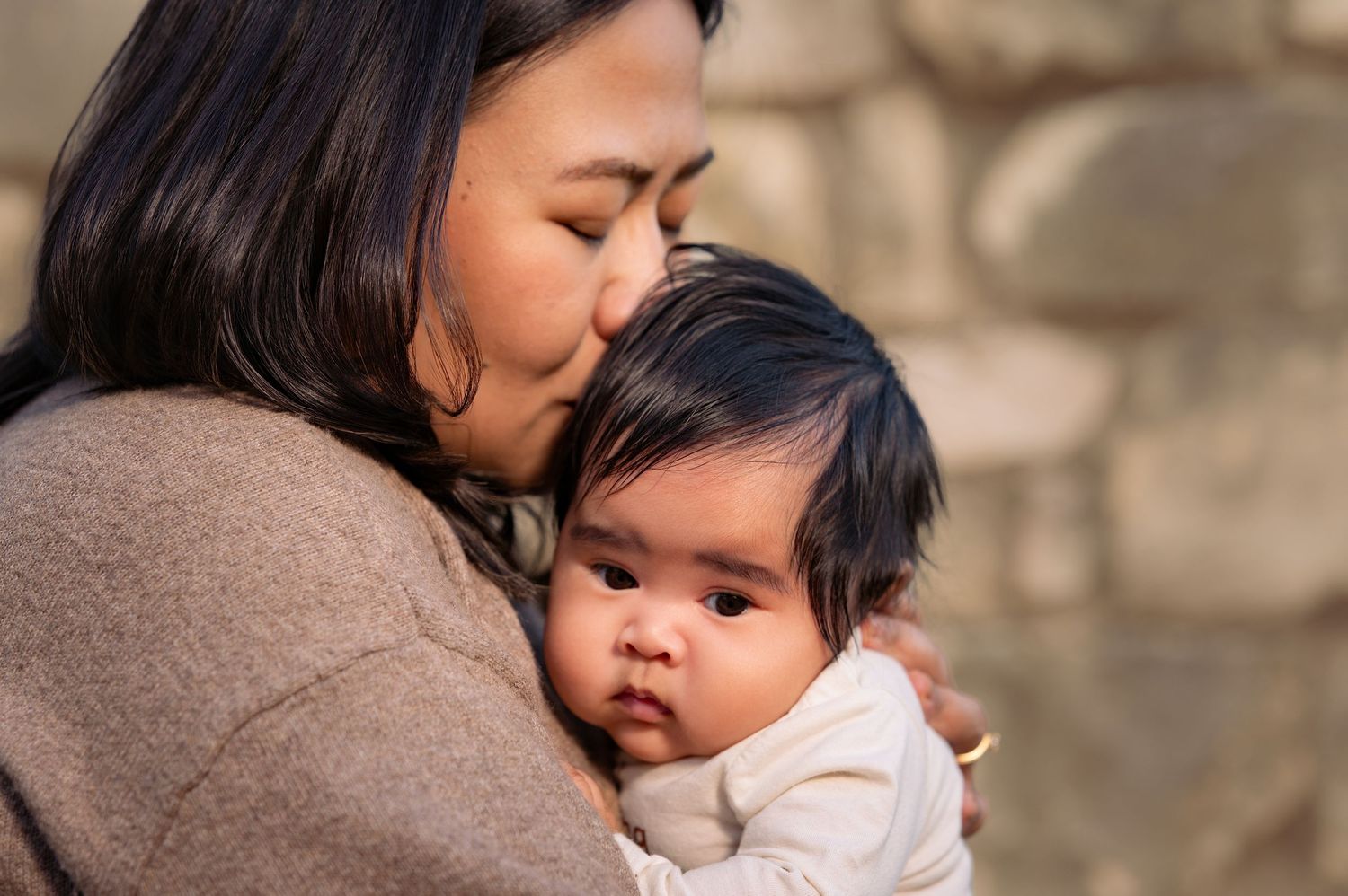 A tender moment between parent and baby as they embrace in warm sunlight against a stone wall.