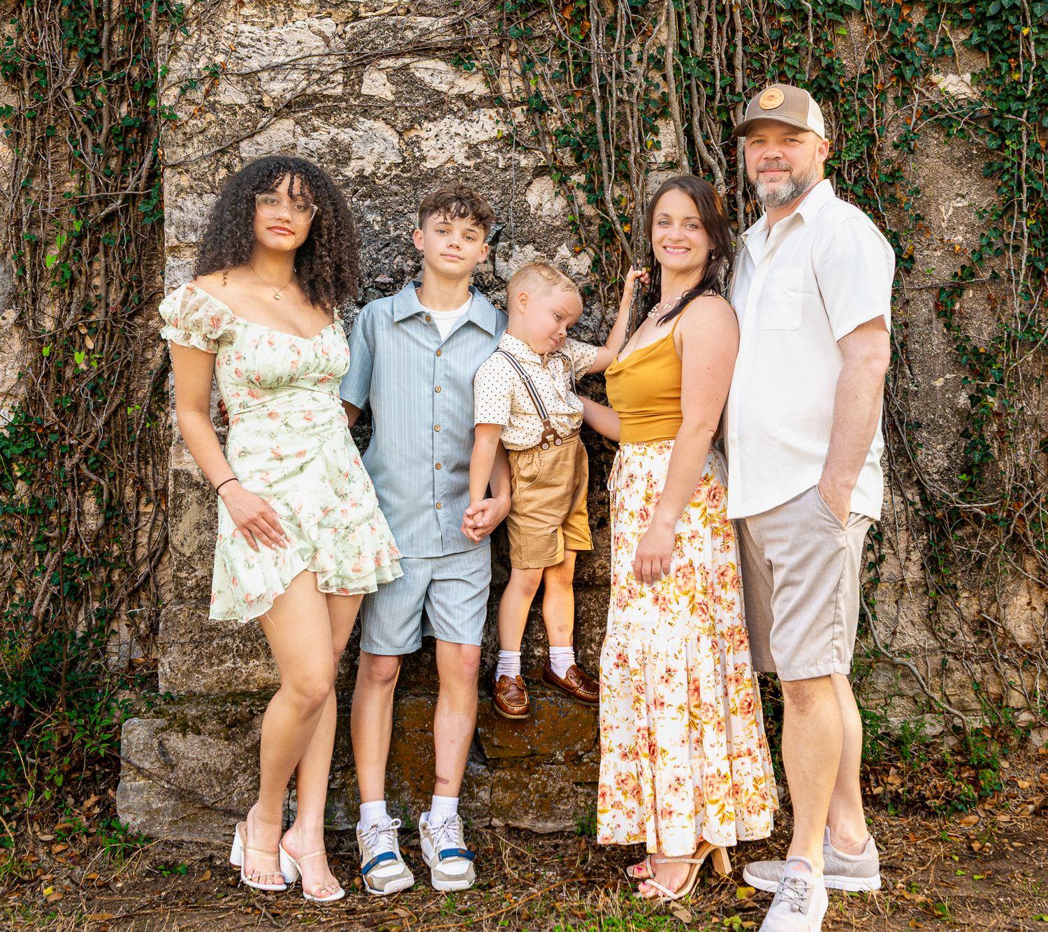 Family group poses together against stone wall backdrop in coordinated casual spring outfits.