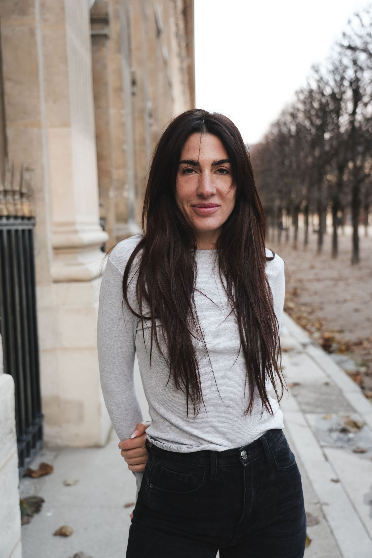A young woman with long dark hair wearing a gray long-sleeve top and black jeans stands on a Parisian sidewalk.
