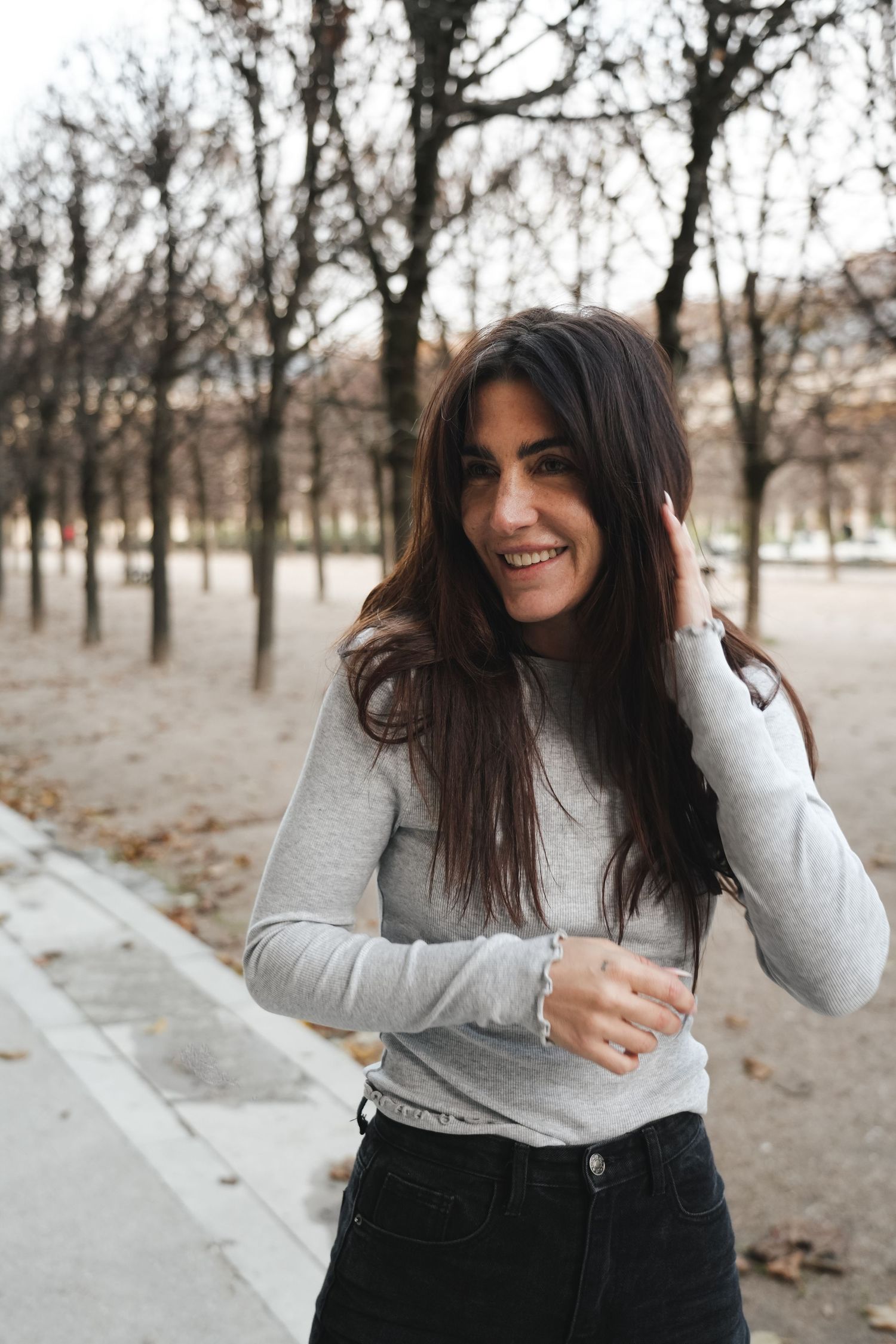Smiling brunette woman in gray long-sleeve top touching her hair while standing in autumn park.