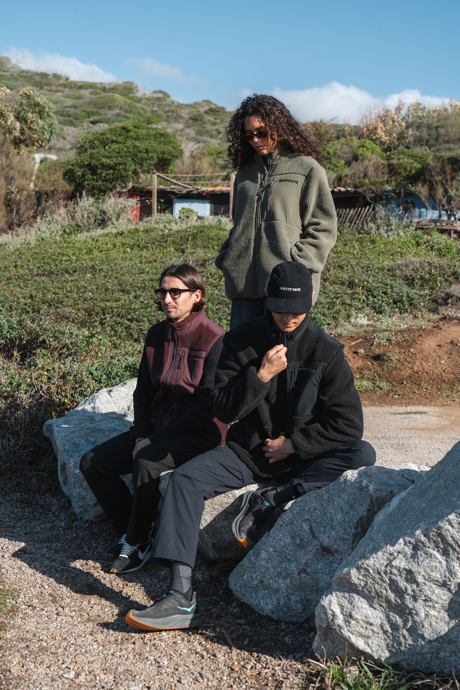 Three friends posing outdoors on rocks wearing casual streetwear including fleece jackets and a black cap.