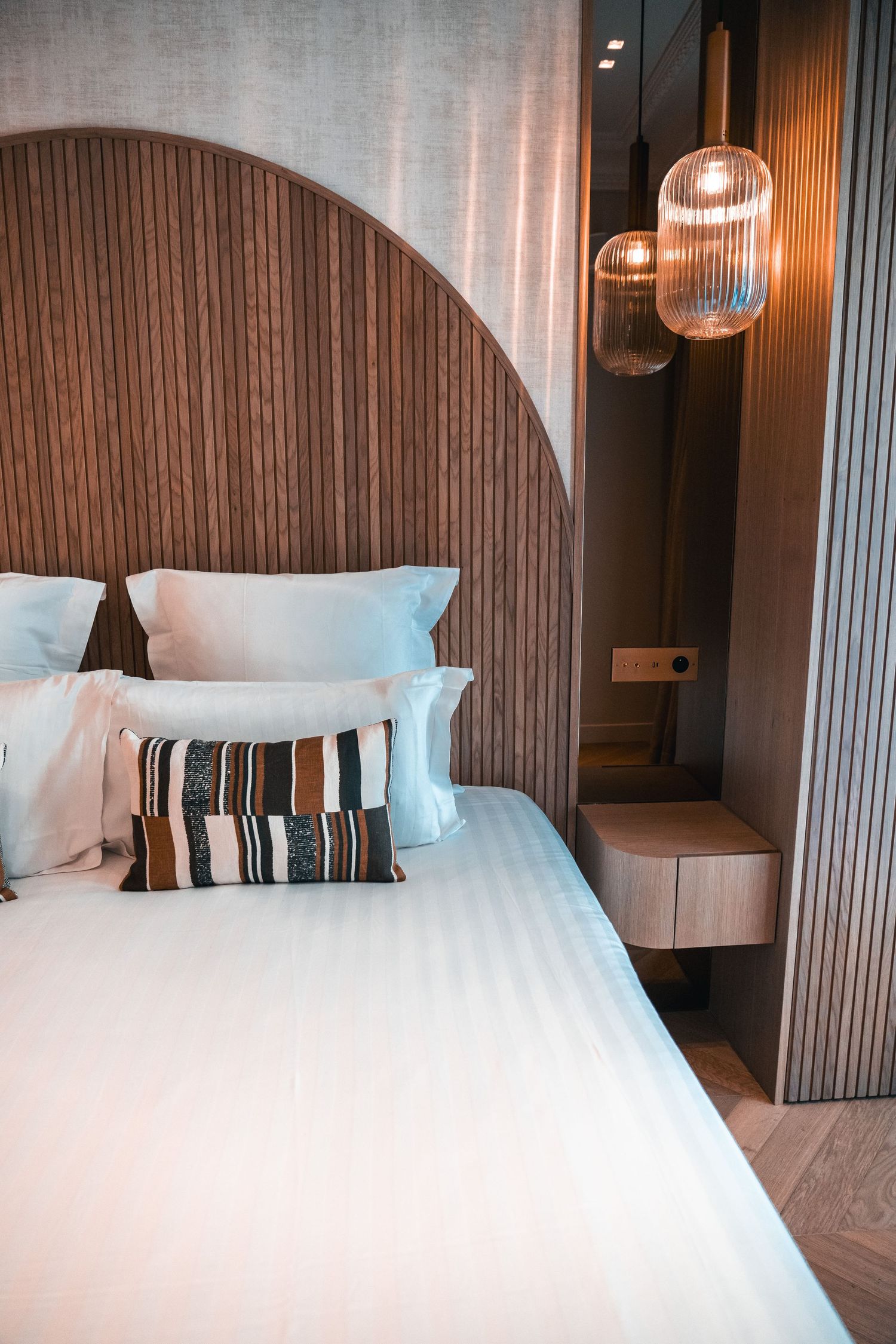 Modern hotel bedroom with arched wooden headboard, white bedding, striped accent pillow, and warm pendant lights.