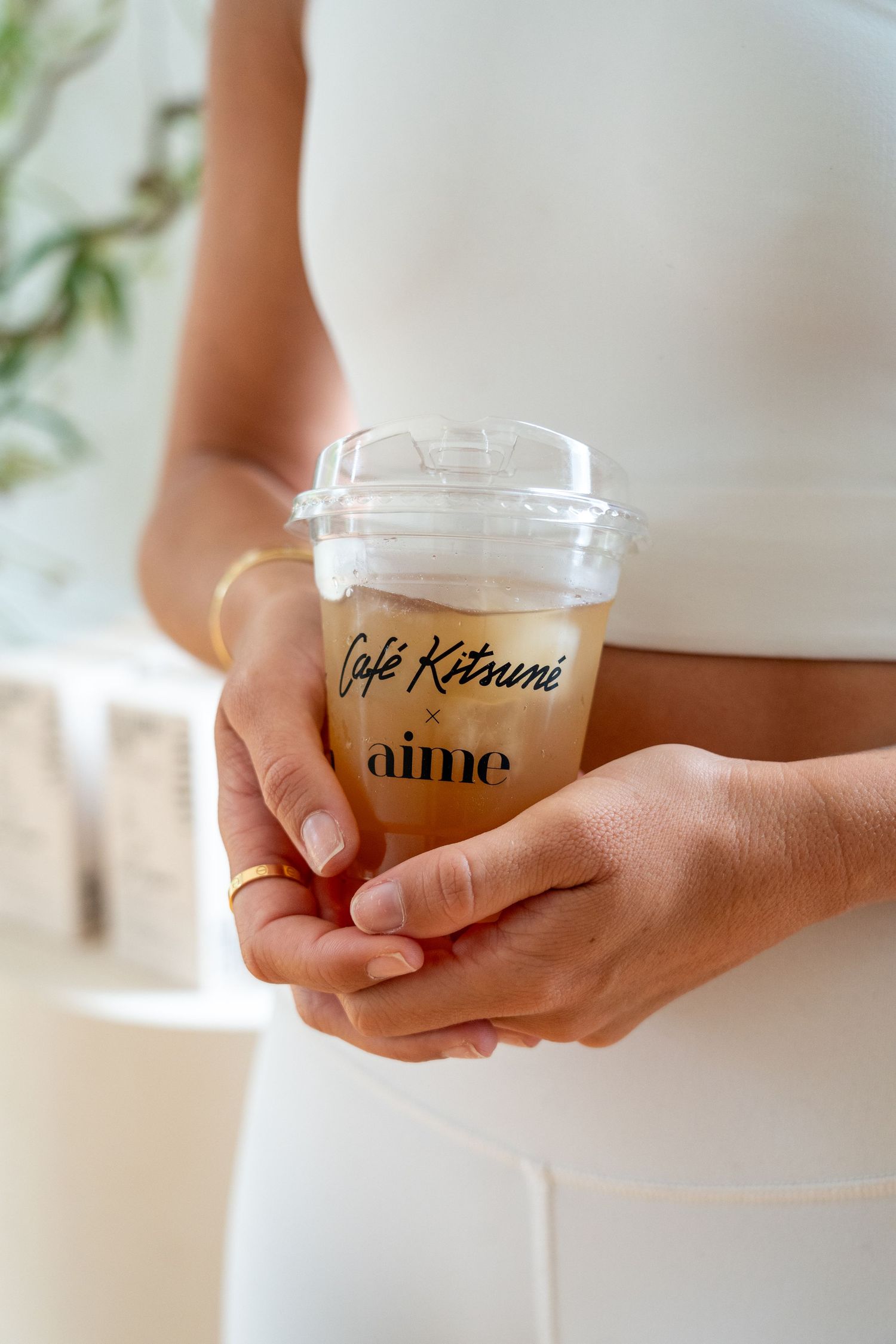 Woman in white outfit holding an iced coffee cup branded with Café Kitsuné x Aime collaboration.