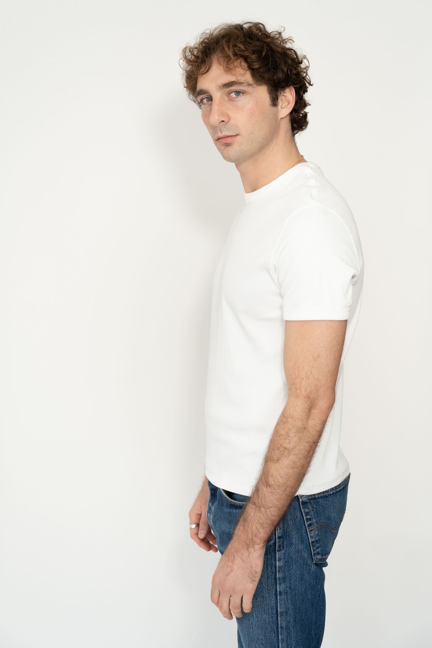 Man in white crew neck t-shirt and blue jeans posing sideways against white background.