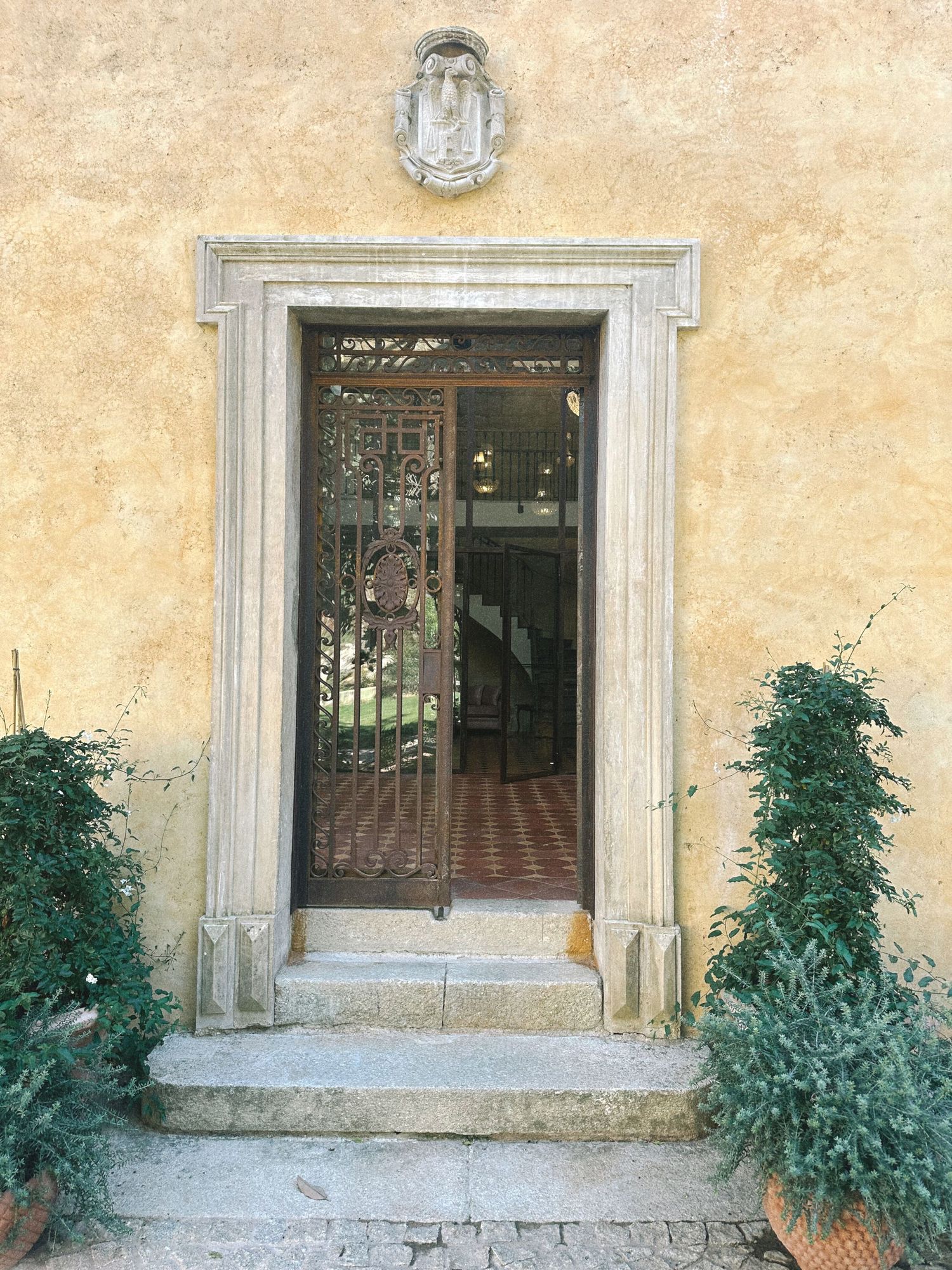 Elegant Italian villa entrance with ornate iron gate, marble doorframe, flanking potted shrubs, and decorative relief above.