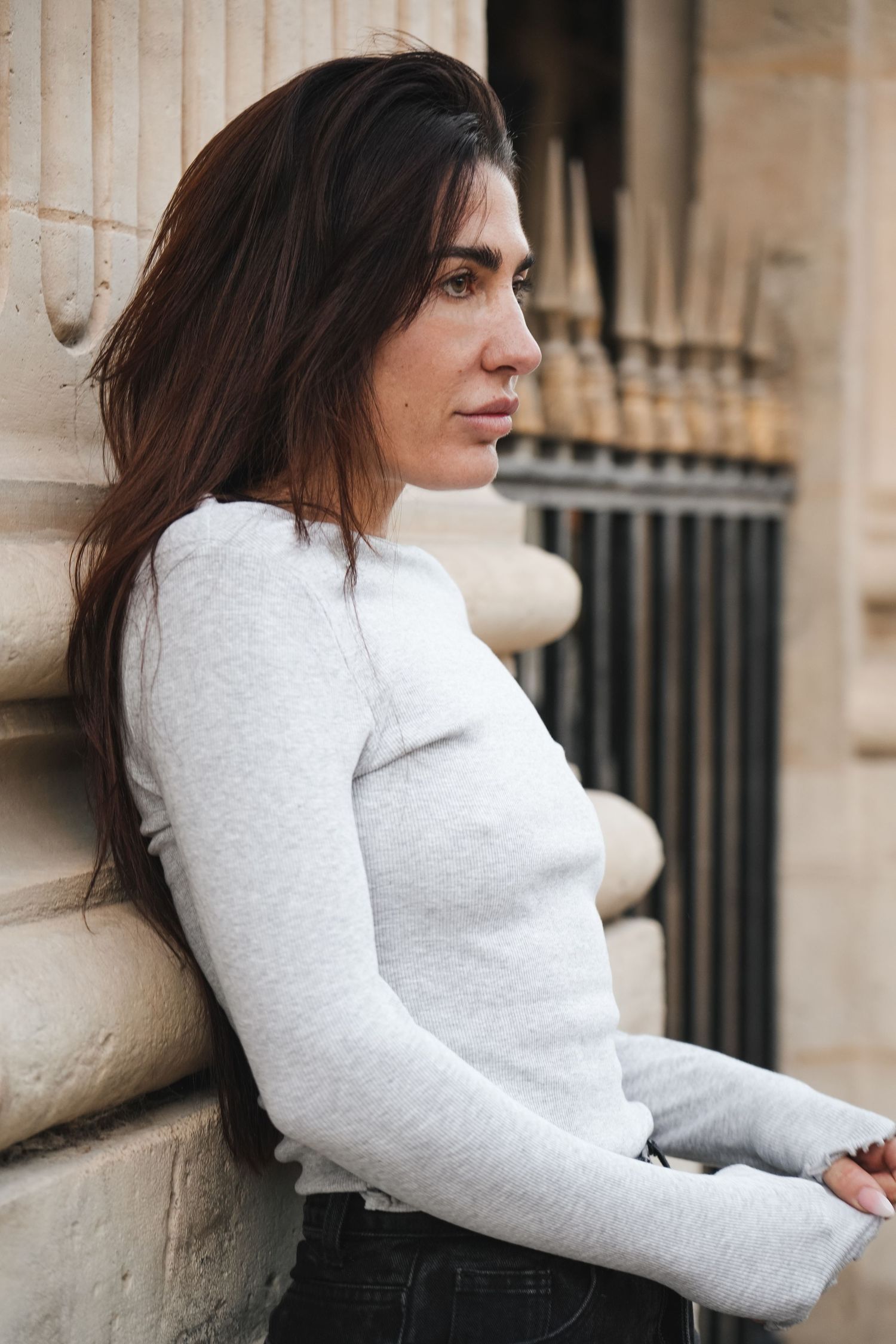 A young woman with long dark hair leans against a stone column wearing a light gray long-sleeve top and dark jeans.