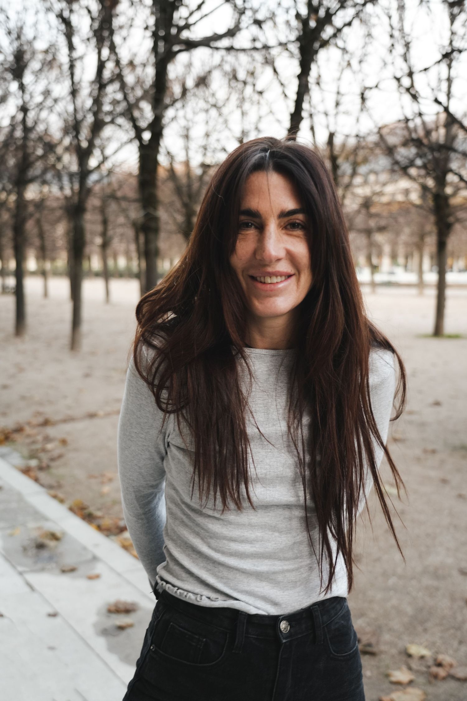 A smiling woman with long dark hair wearing a gray top and black jeans standing in an autumn park.