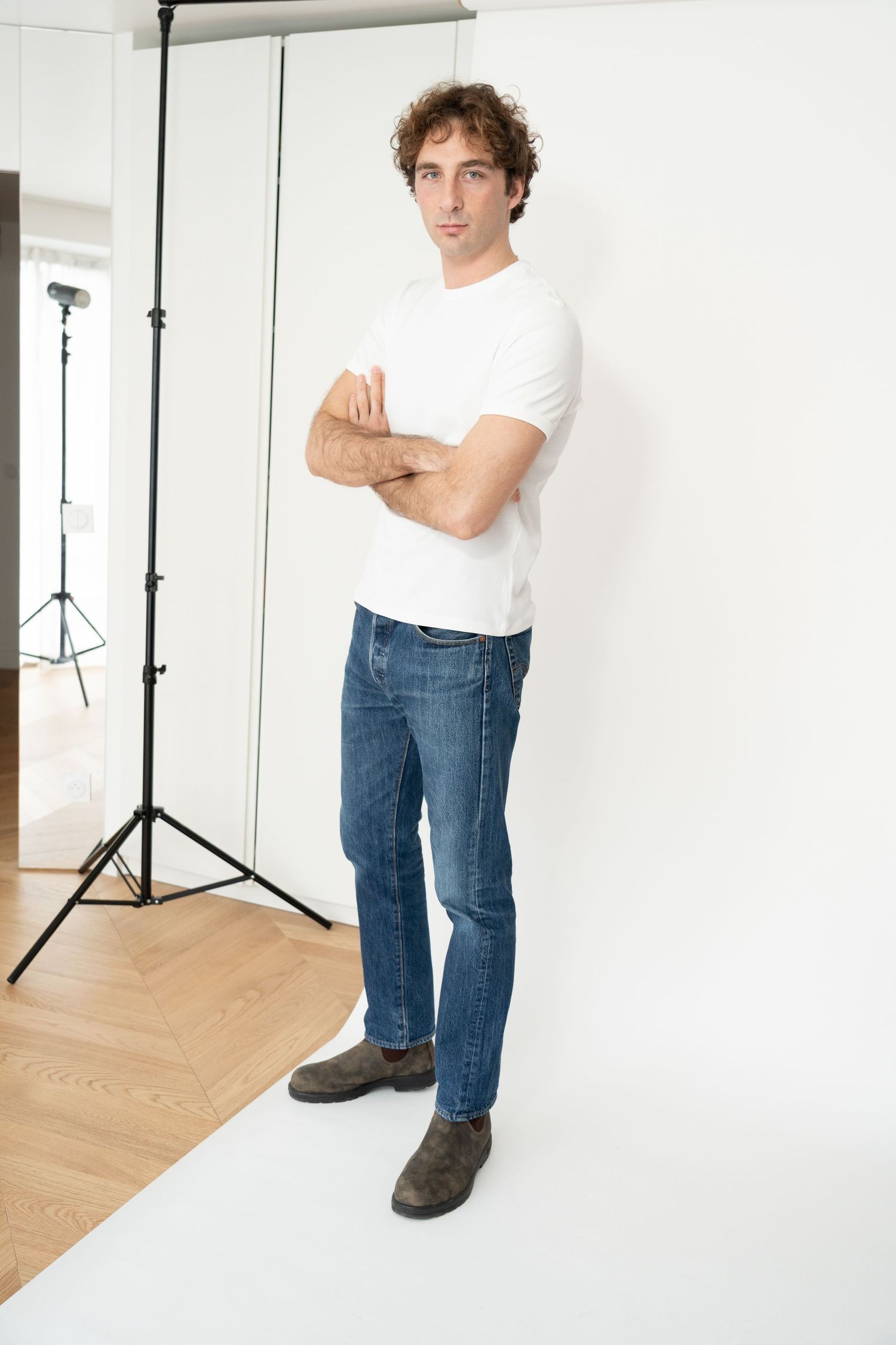 Man in white t-shirt and blue jeans standing with arms crossed in a bright studio setting.