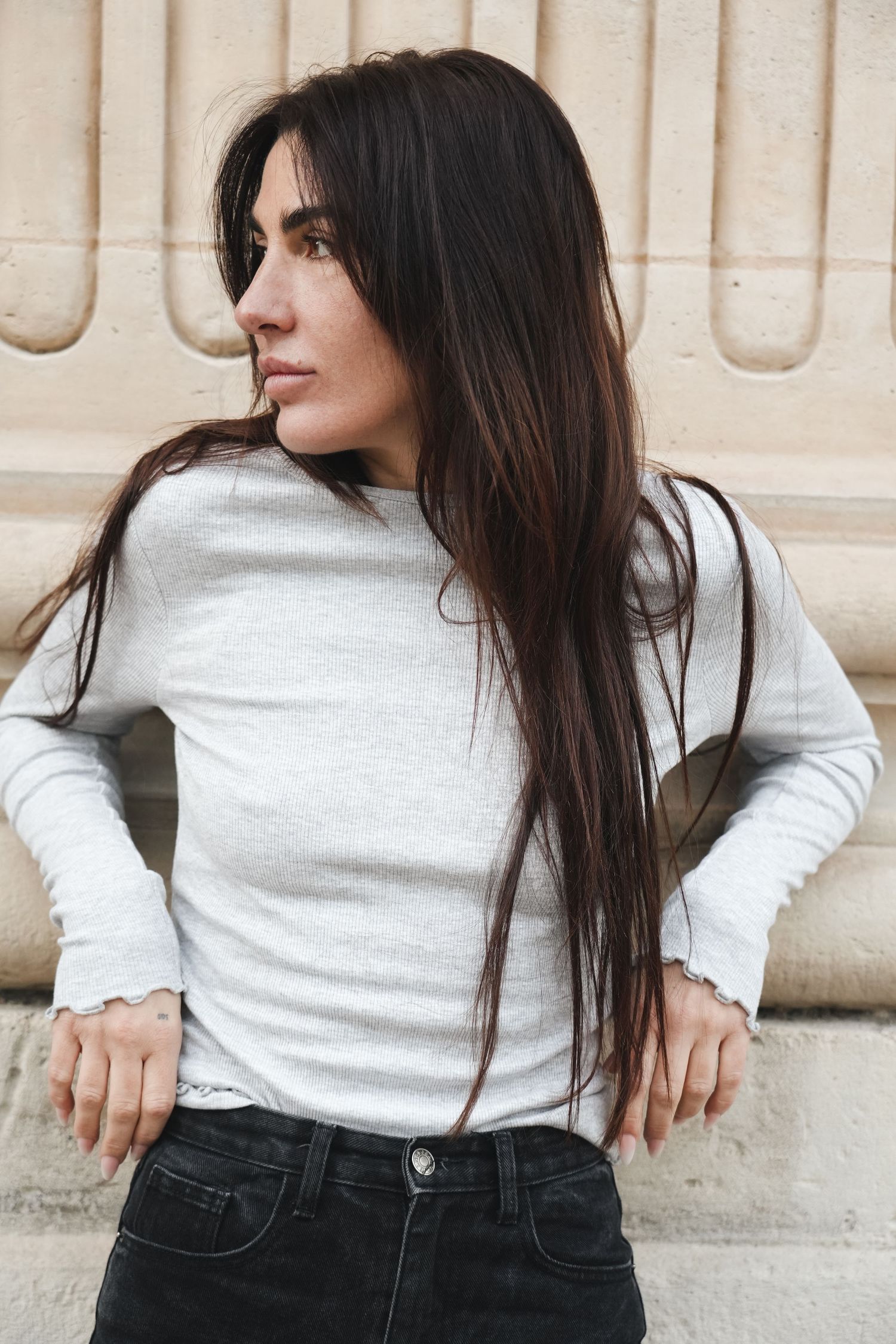 Young woman with long dark hair in gray long-sleeve top and black jeans posing against beige wall outdoors.