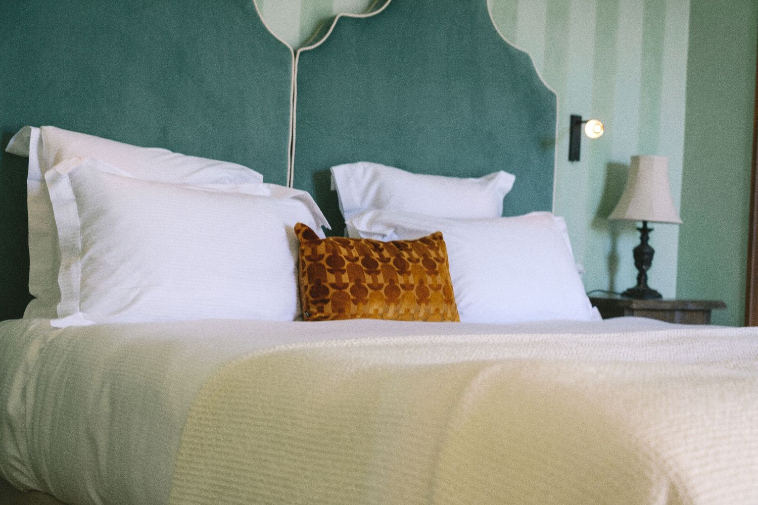 Elegant hotel bed with white pillows, cream bedding, and a gold velvet accent pillow against a teal upholstered headboard.