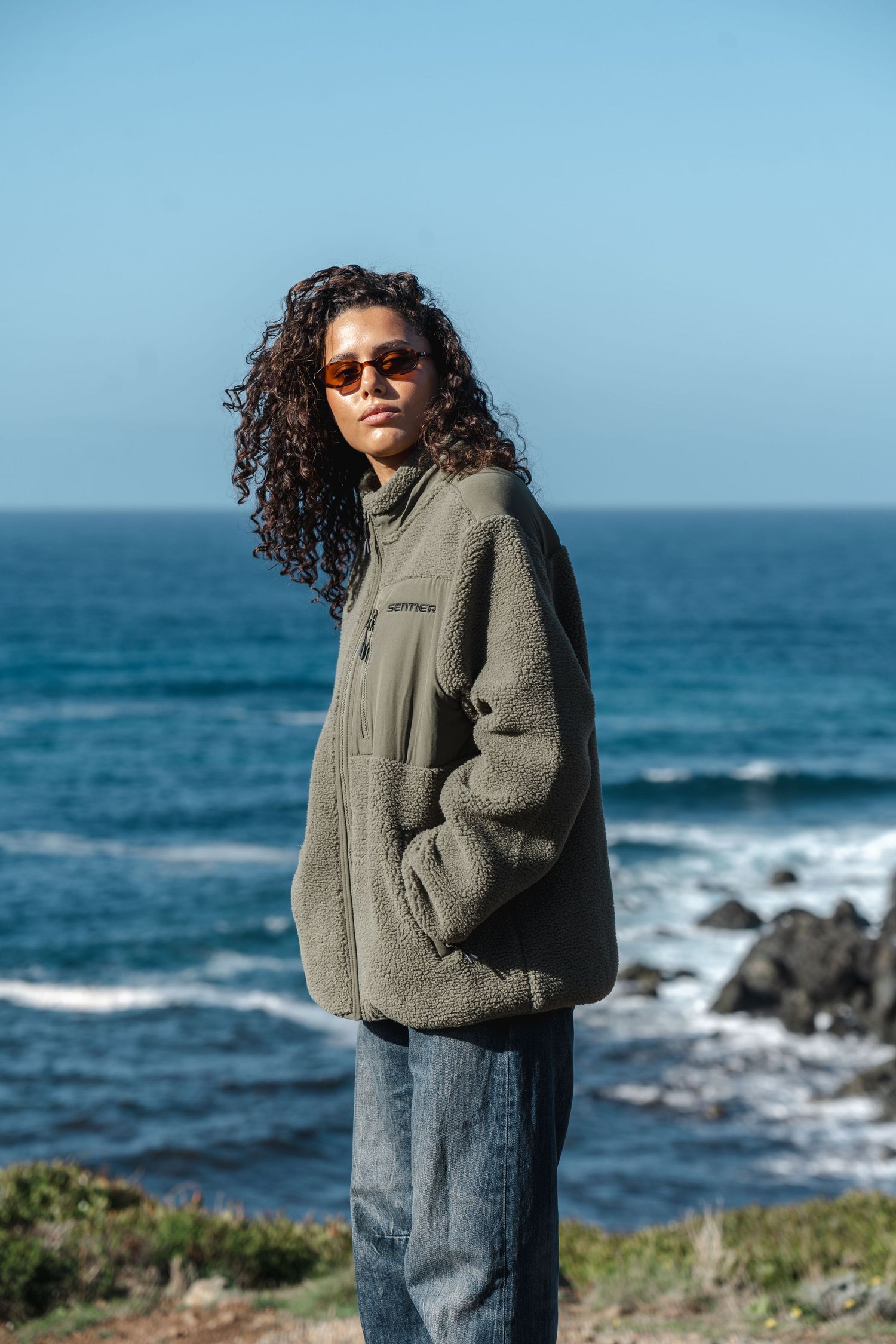 Person with curly hair wearing sunglasses and olive fleece jacket standing by rocky ocean coastline on clear day.