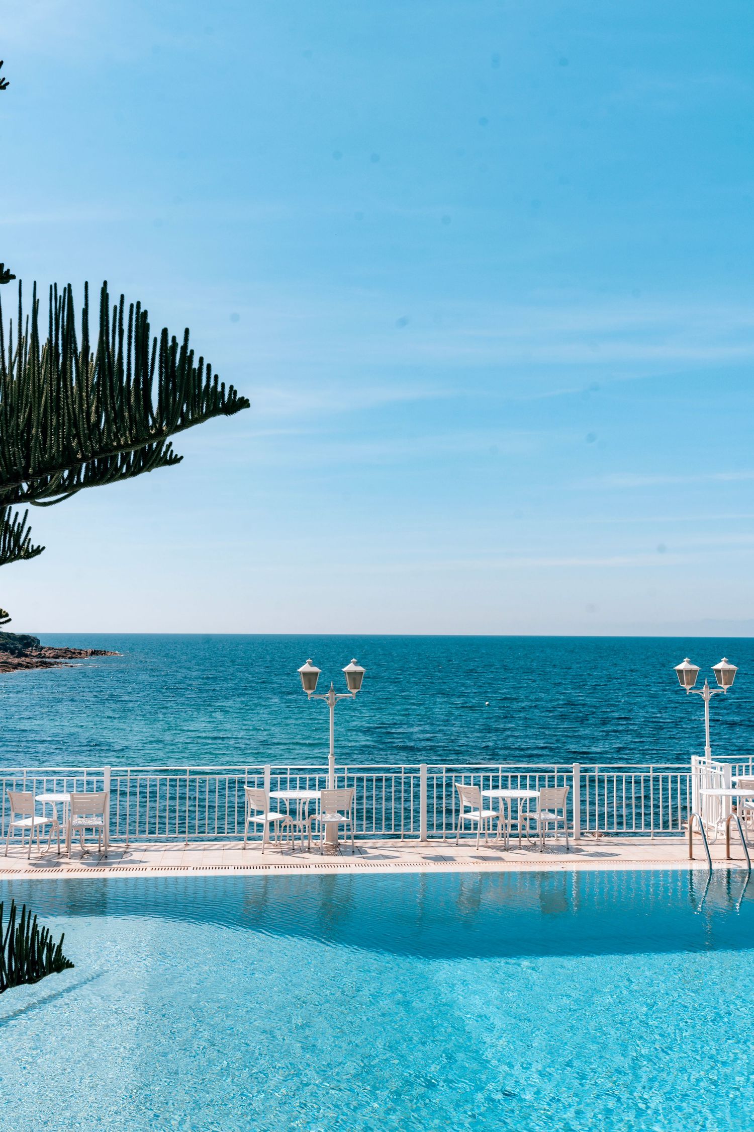 Luxury resort infinity pool overlooking turquoise ocean with white railing, outdoor dining, and tropical plants under blue sky.