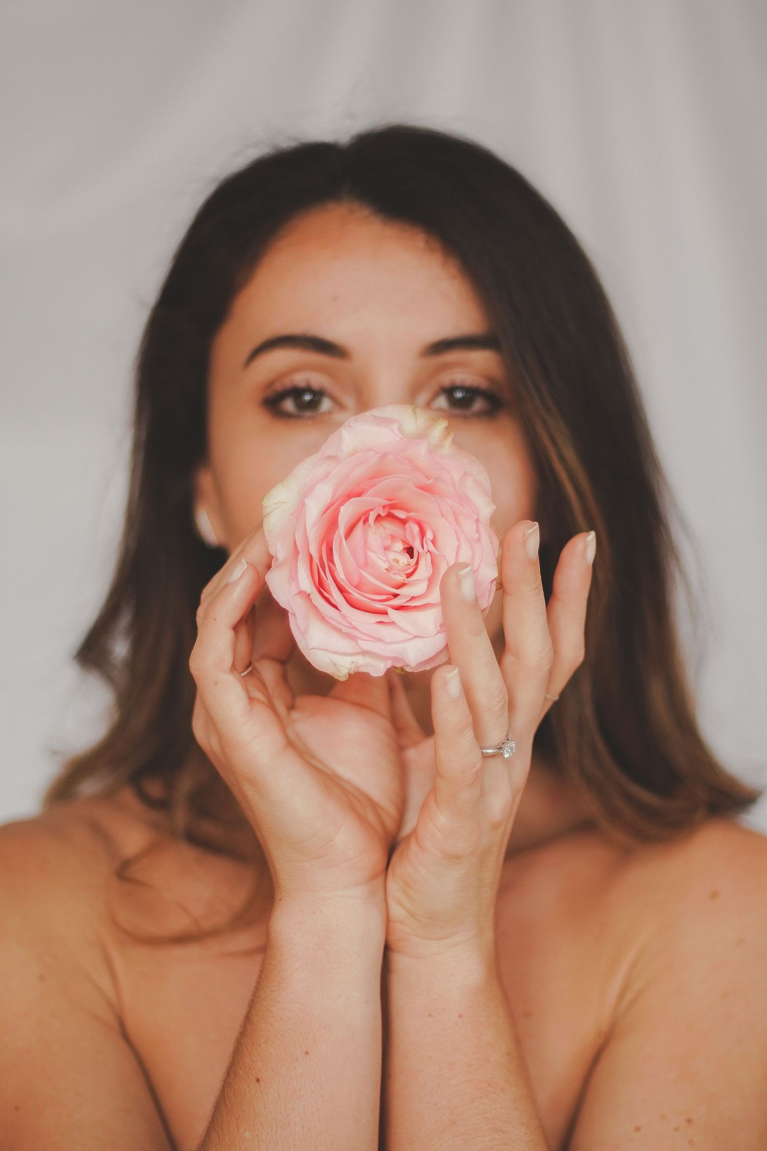 Woman holding a pink rose in front of her face with both hands, wearing a silver ring, against a white background.